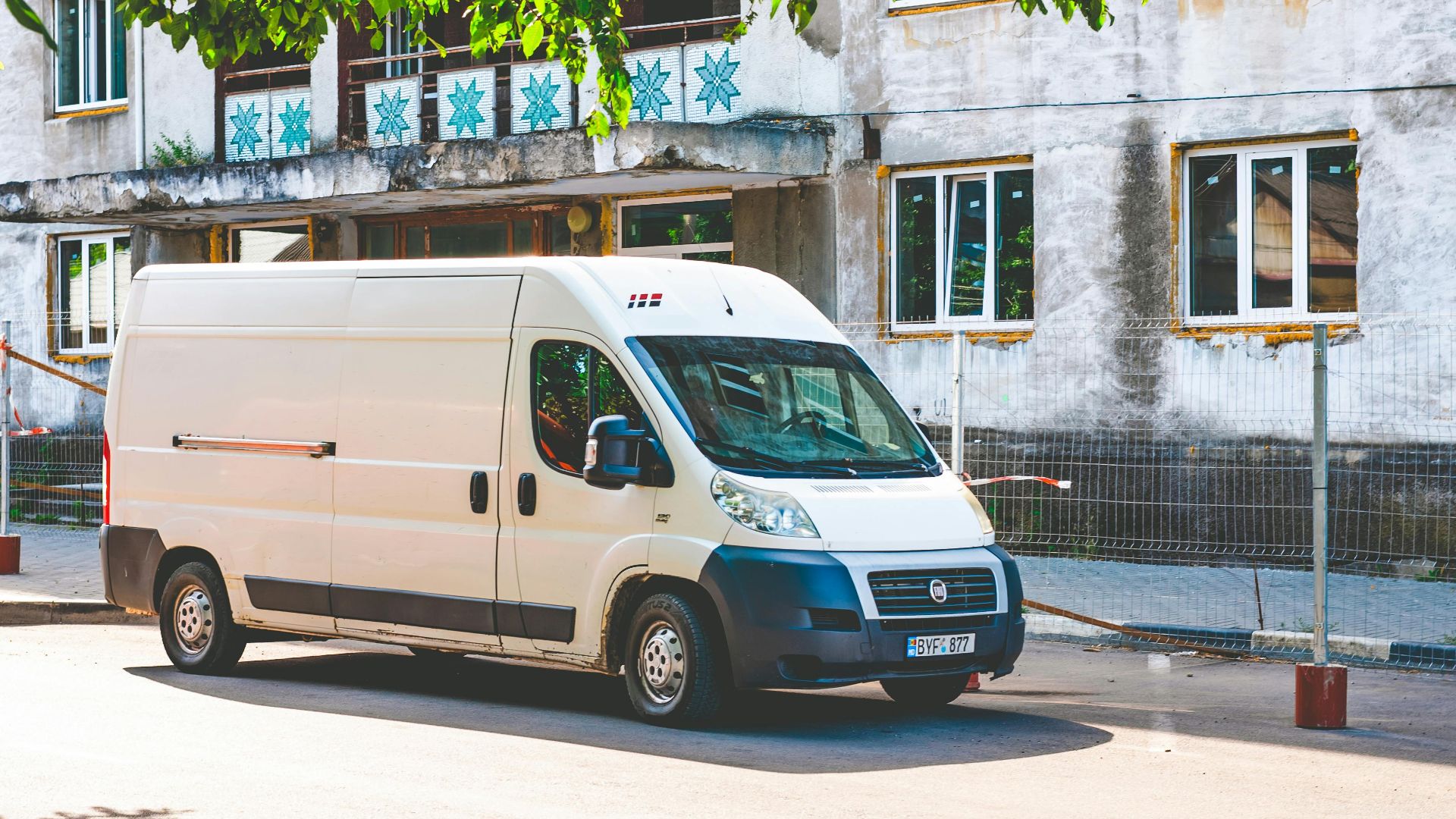 A white delivery van parked under a tree on a sunny day in an urban area.