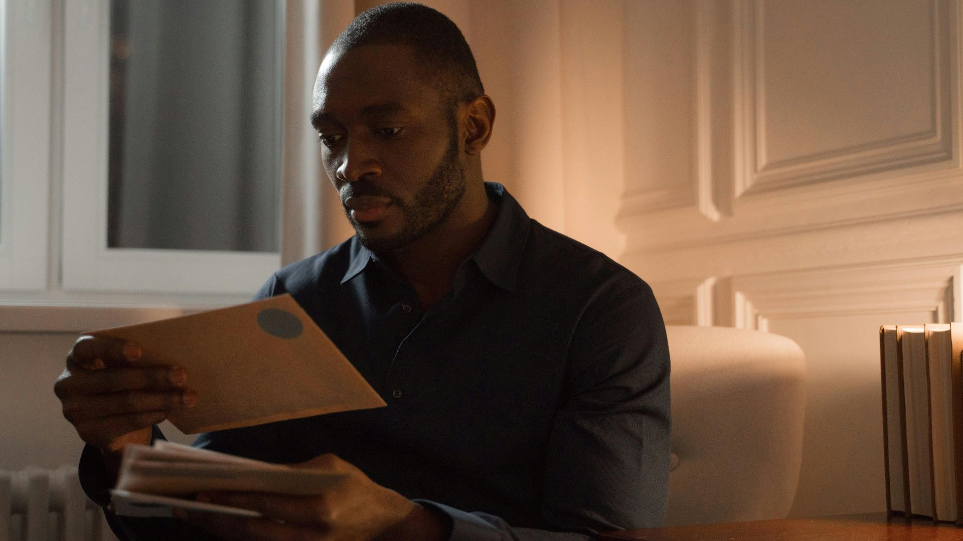 A man with a mustache reads letters while sitting in a warmly lit home interior.