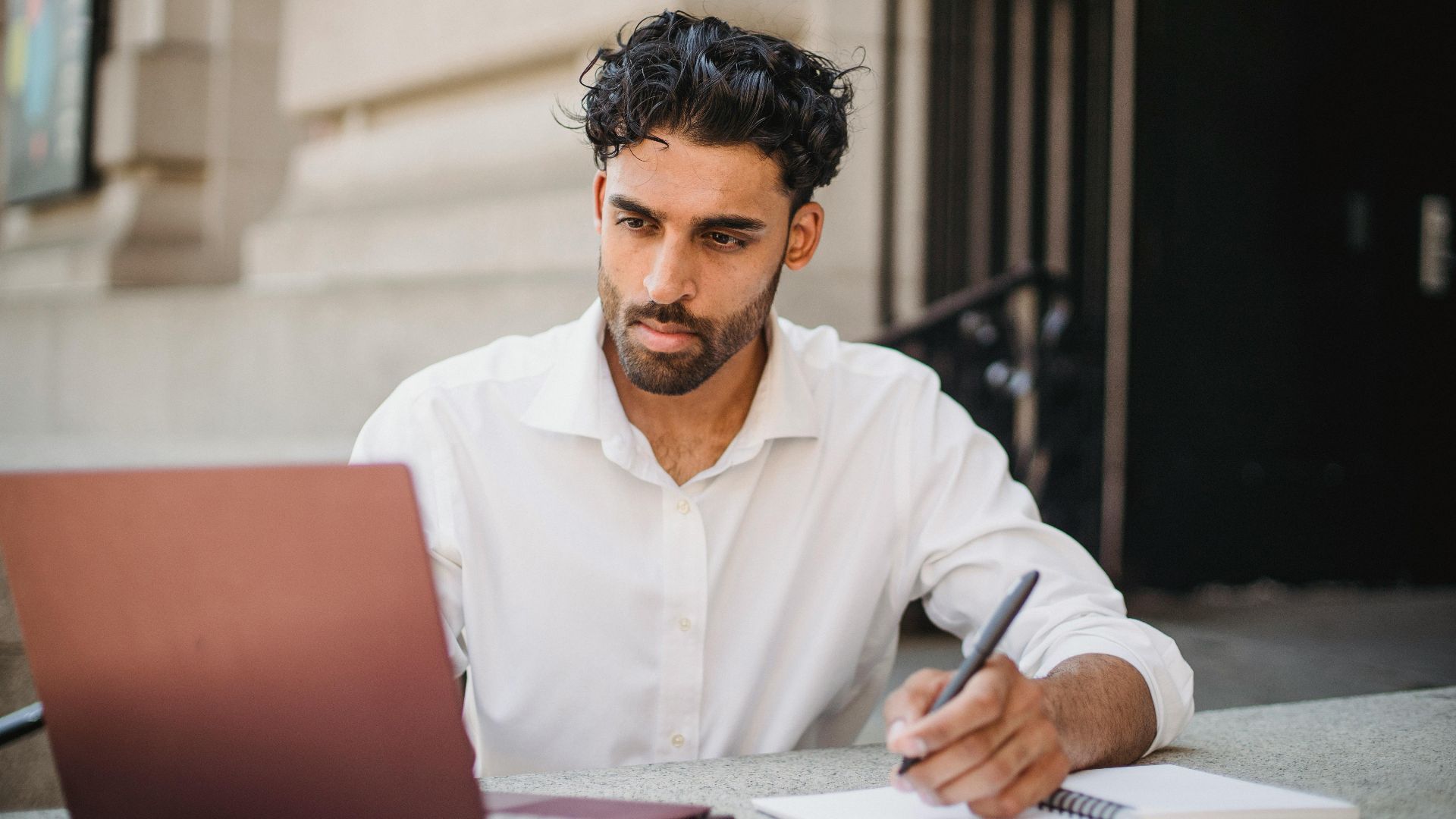 Focused businessman working on laptop outdoors, taking notes in notepad, wearing formal white shirt.