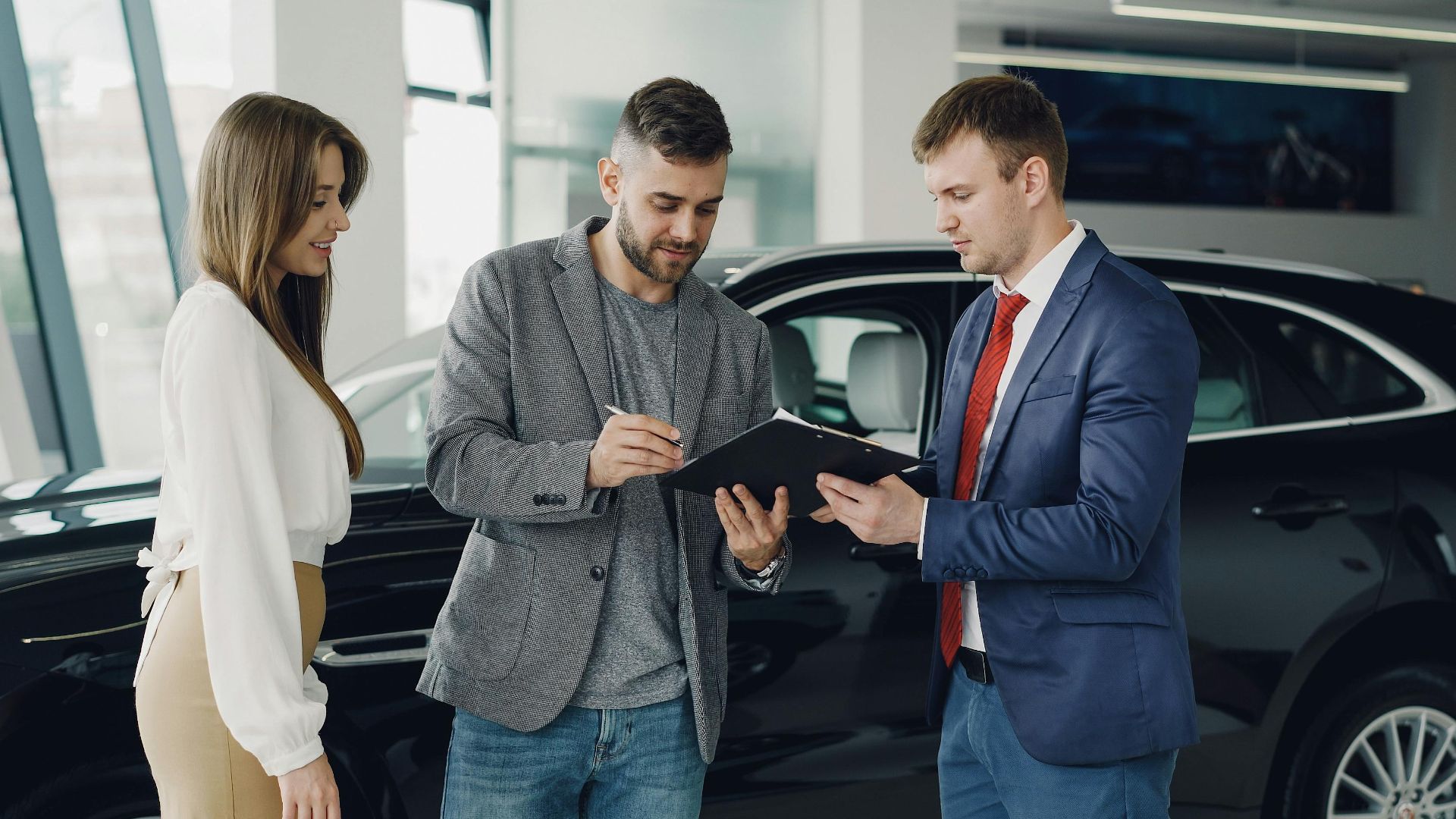 Three adults discussing documents at a car dealership beside a black car.