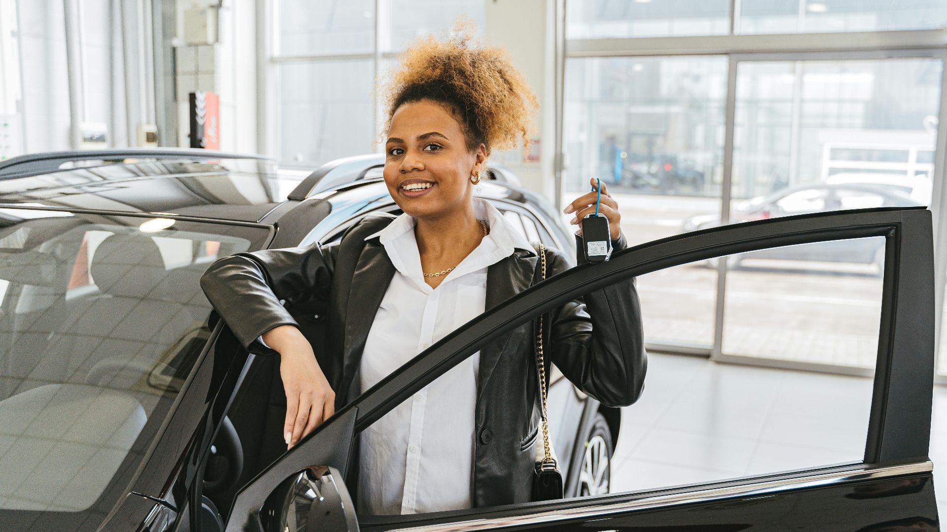 Smiling woman holding car key in dealership showroom next to new vehicle.