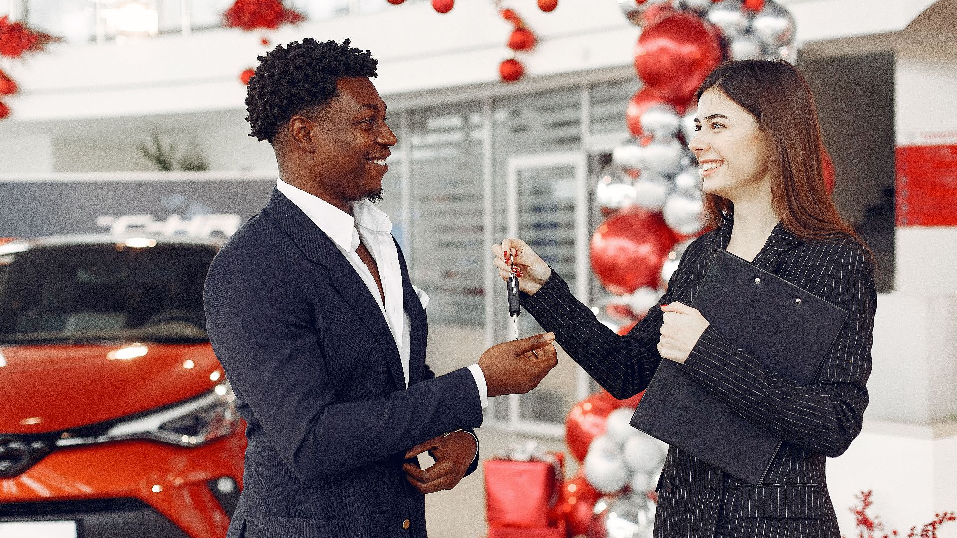Side view of happy positive female dealer in formal classy wear passing car keys to  excited African American customer in trendy suit in car showroom