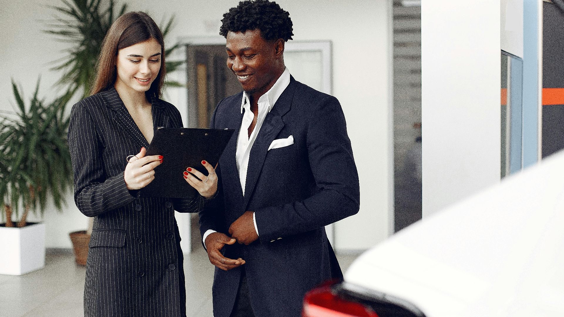 Cheerful multiethnic stylish female agent and smiling customer in formal suit discussing contract details while standing in modern car showroom in daylight