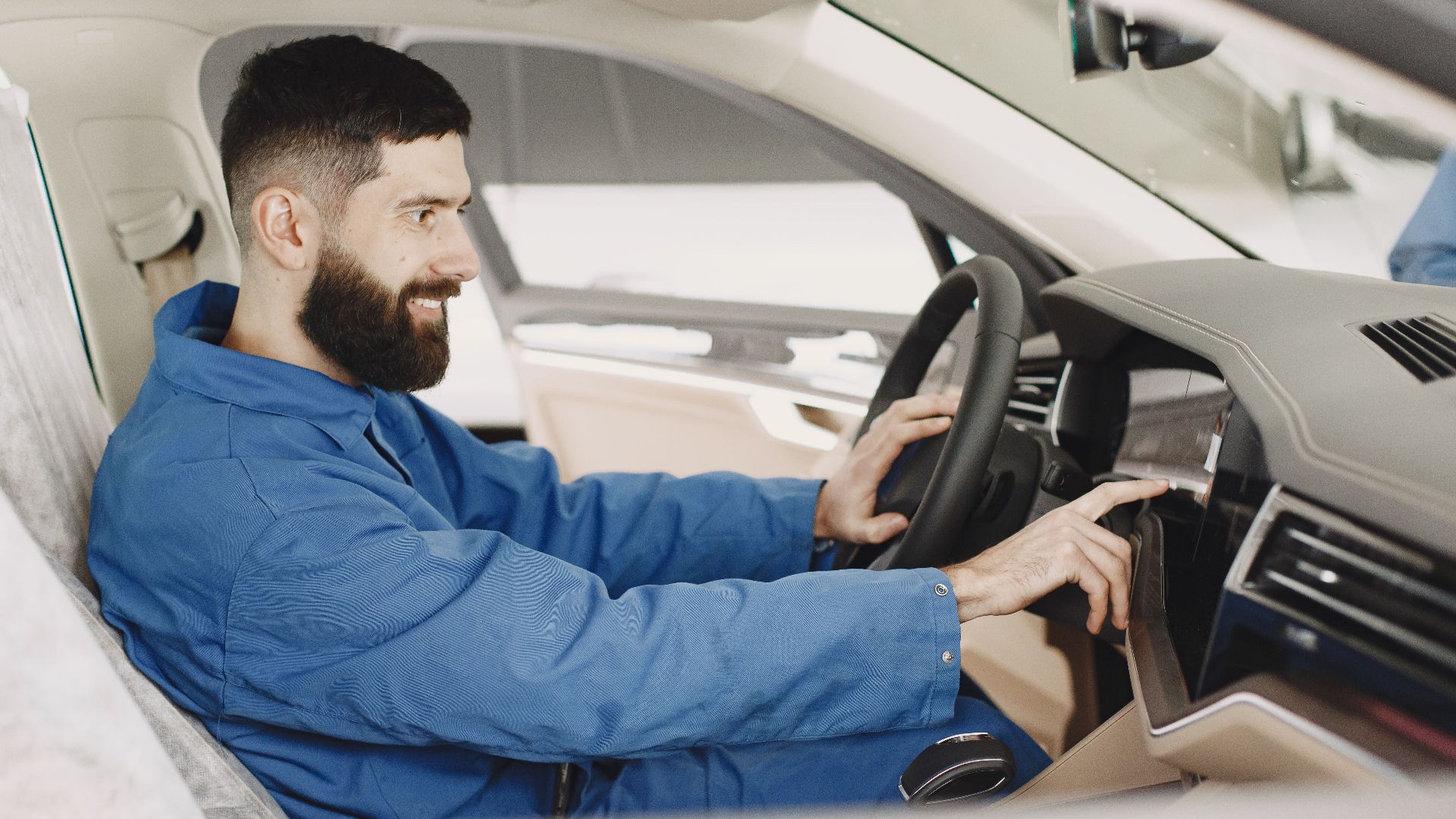 Mechanic in blue coveralls interacts with car dashboard, smiling and focused.