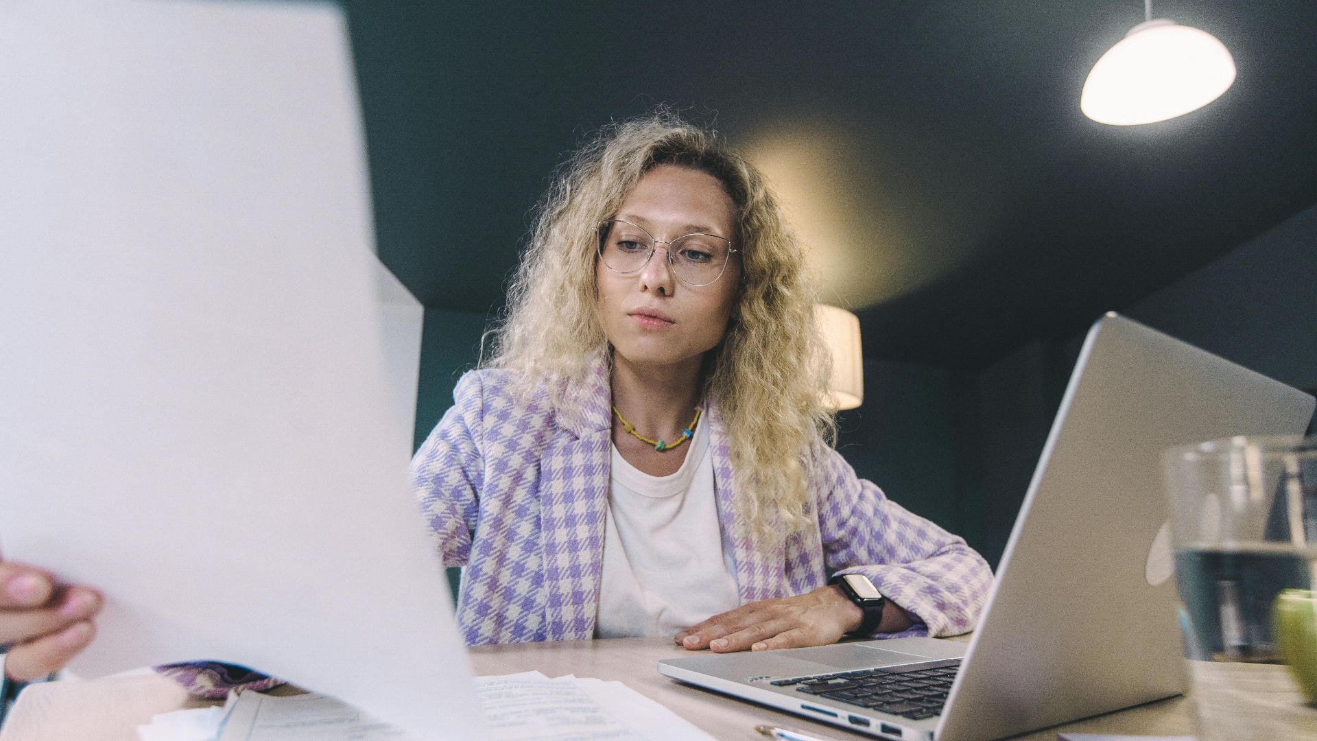 Woman in an office environment reviewing documents with focus, surrounded by technology.