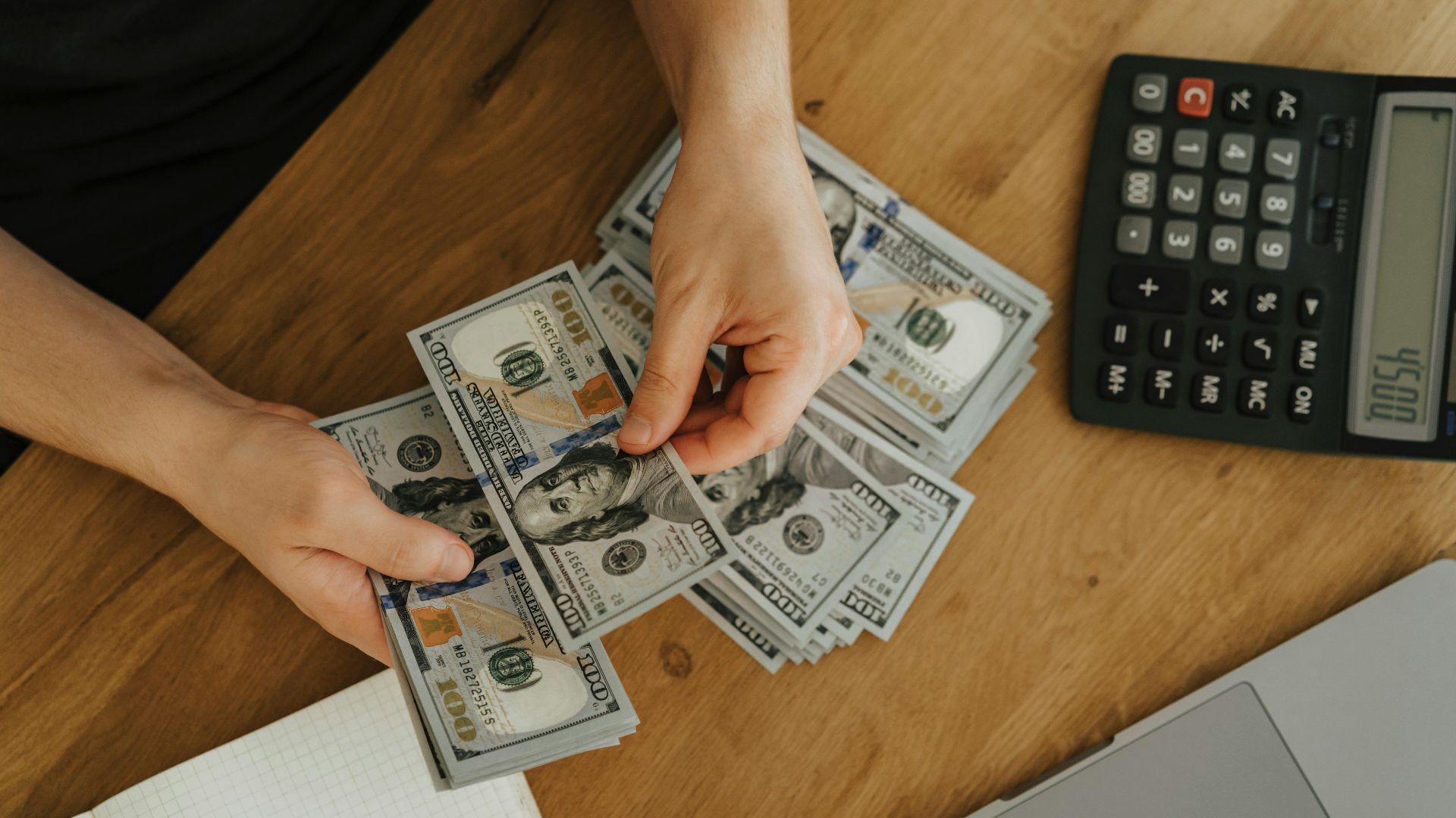 Close-up of hands counting dollar bills with calculator, notebook, and laptop on wooden table.