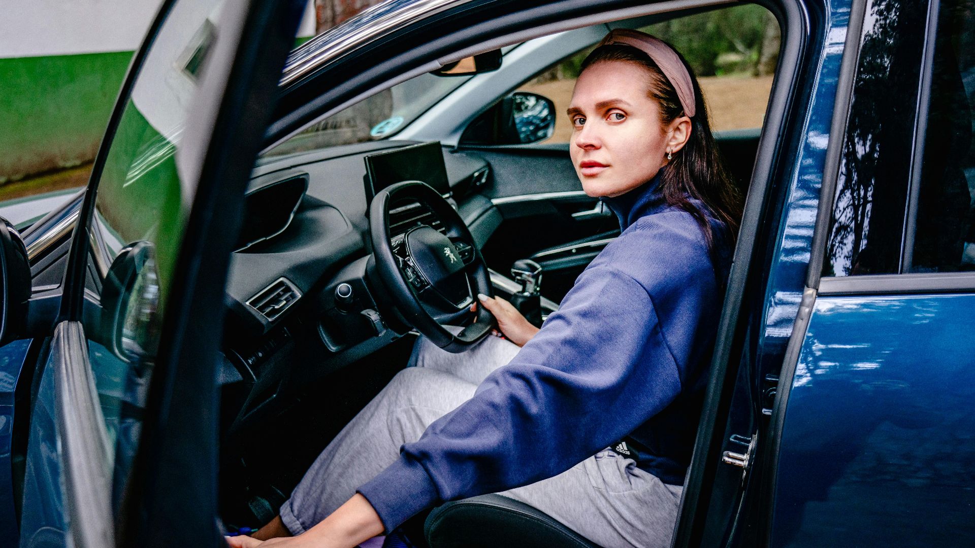 Woman sitting in the driver's seat of a car with an open door, looking outside. Natural setting.