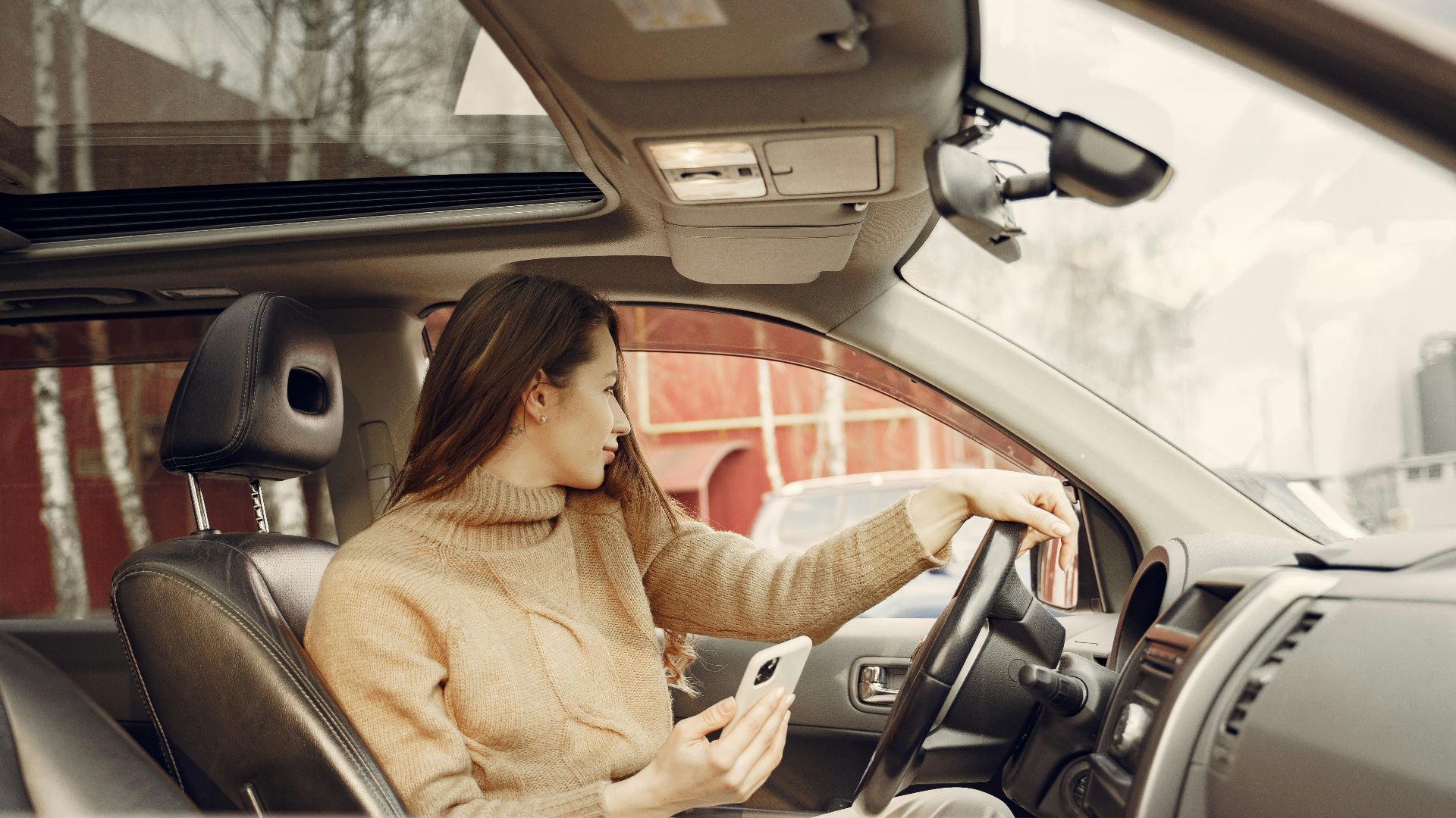 From window side view of adult woman in warm clothes sitting at wheel of modern car waiting for friend and using smartphone