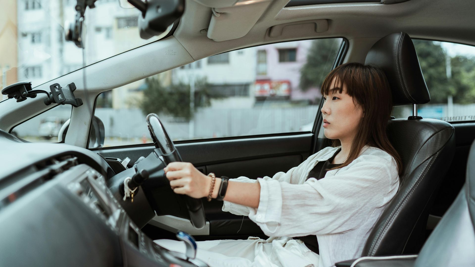 Side view unemotional Asian female in casual white clothes steering right hand drive car along urban streets in daytime