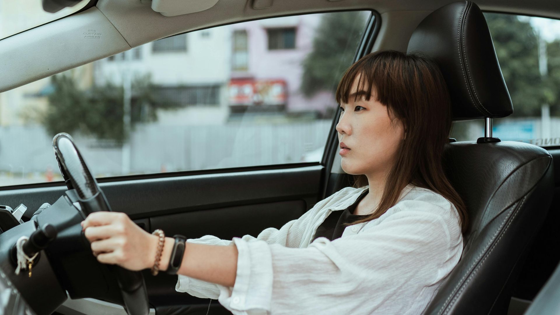 Side view unemotional Asian female in casual white clothes steering right hand drive car along urban streets in daytime