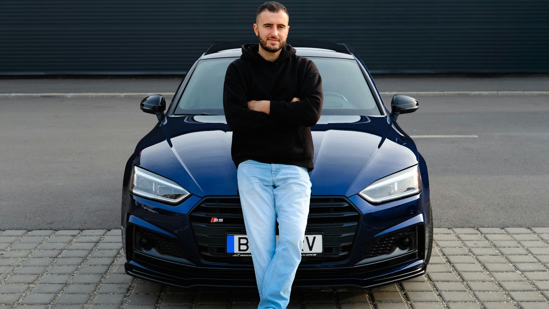 Man casually leaning on a blue car on a street, displaying urban fashion and style.