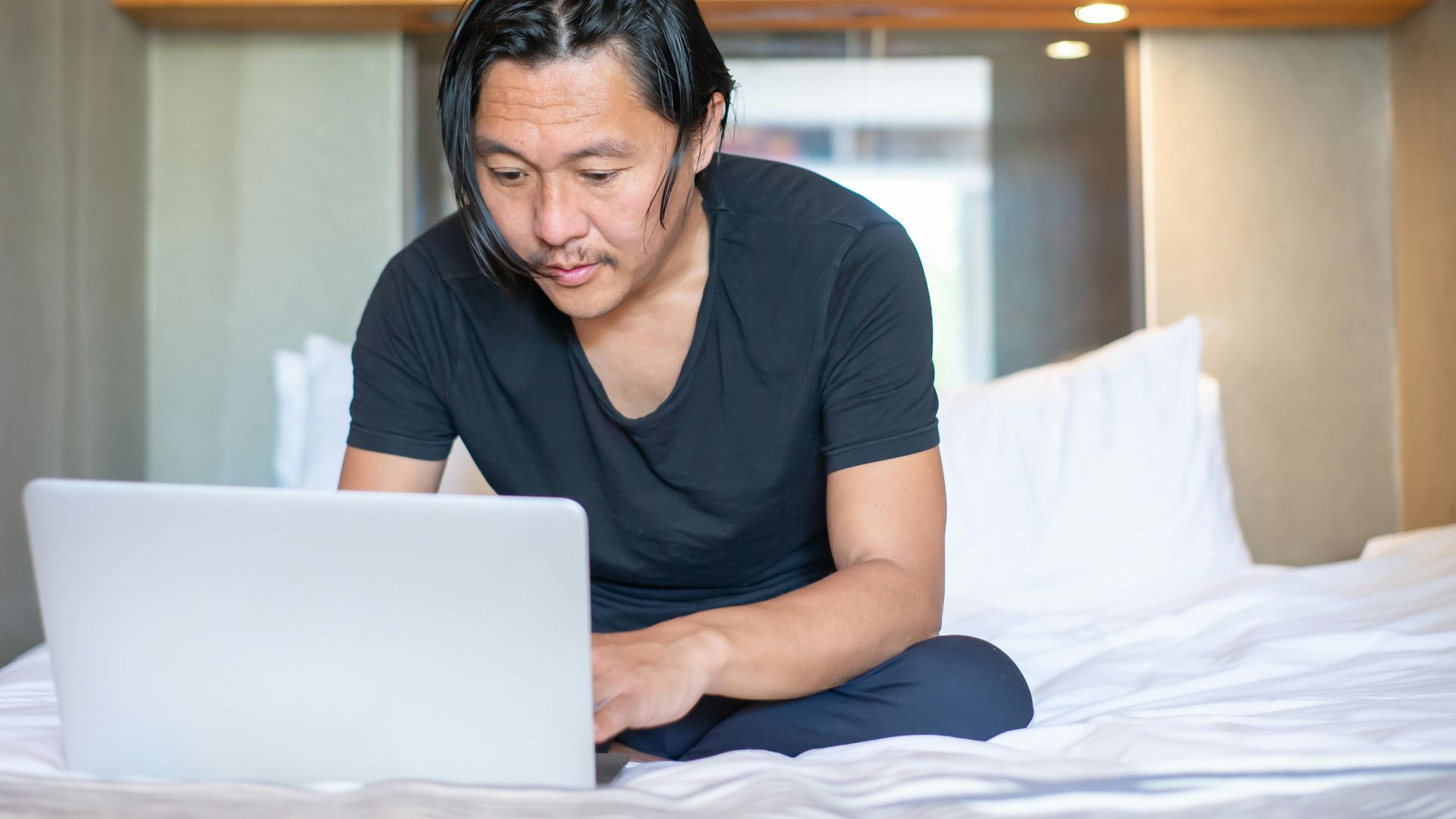 Asian man working remotely on a laptop in a cozy bedroom setting, exemplifying modern work-from-home lifestyle.