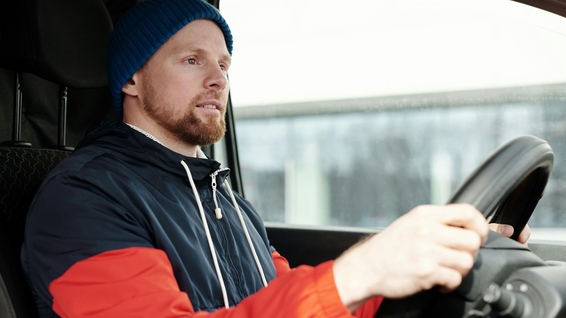 A bearded man wearing a hoodie and knitted cap driving a car, focused and attentive.