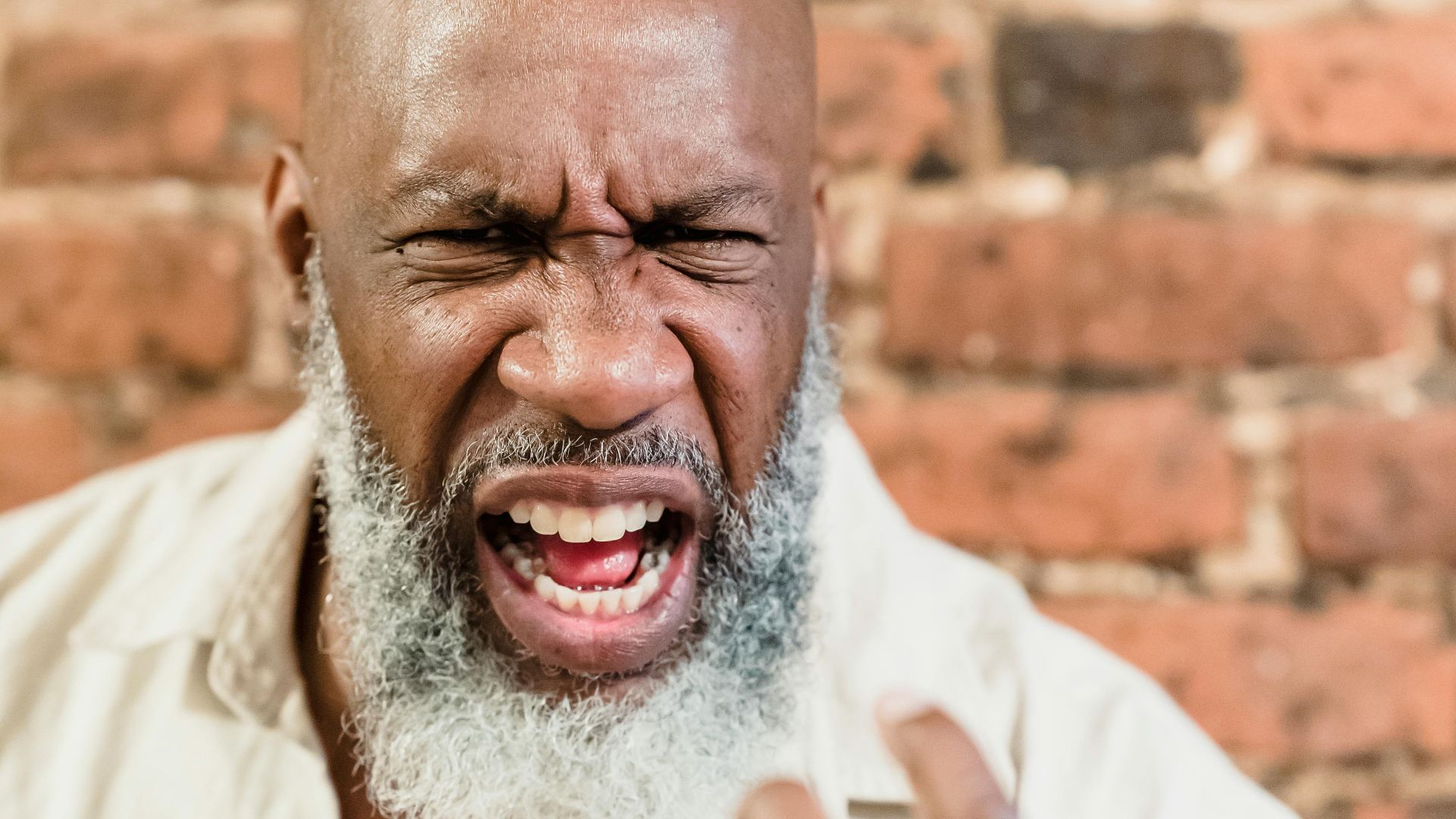 A dramatic close-up of an elderly man with facial hair showing an intense expression against a brick wall.