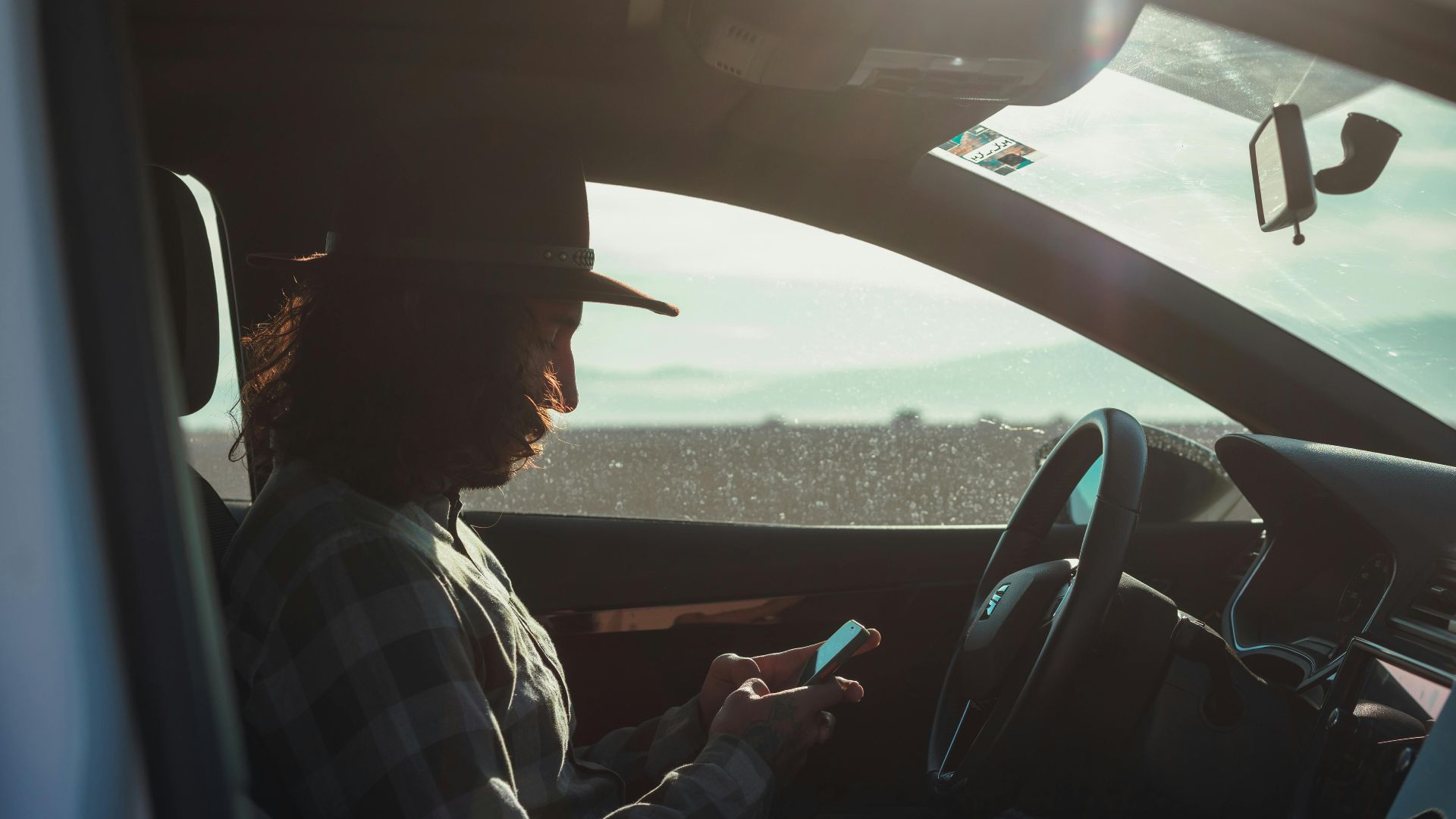 Profile of a man with a hat using a phone inside a car during the day.