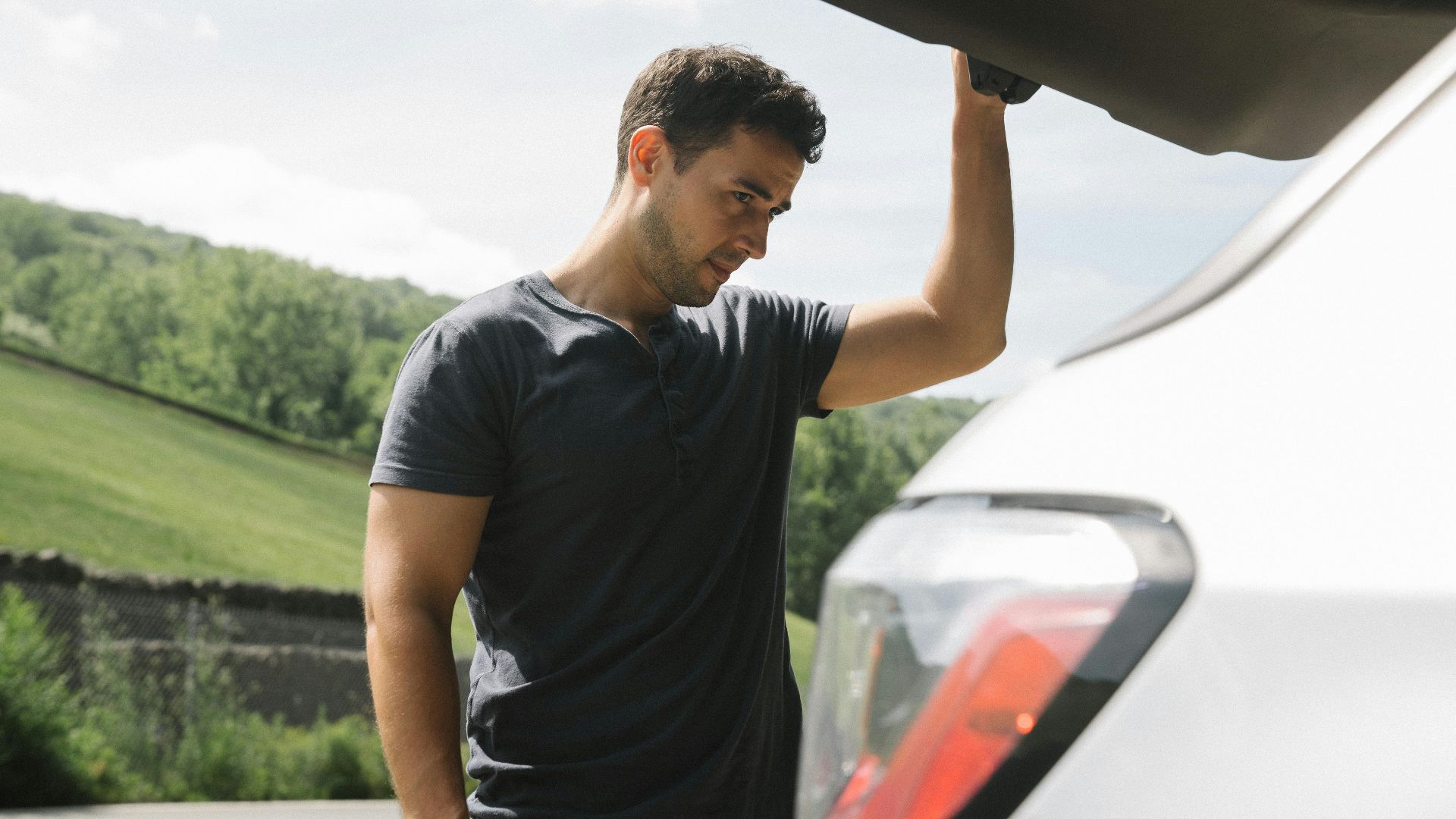 A man examining the open trunk of a car under clear skies with greenery in the background.