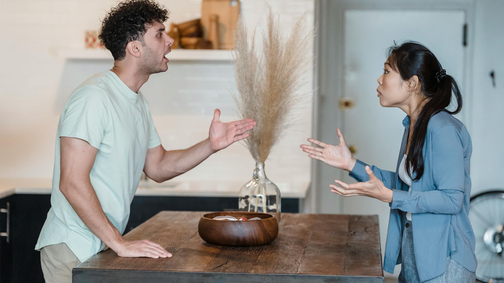 A couple engages in a heated argument at a wooden table in a modern indoor setting.