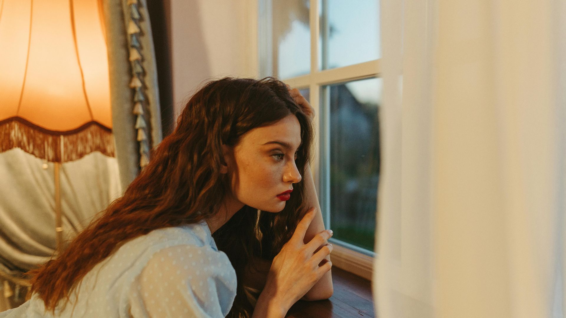 A young woman with red hair gazes thoughtfully out a window indoors, surrounded by warm lighting.