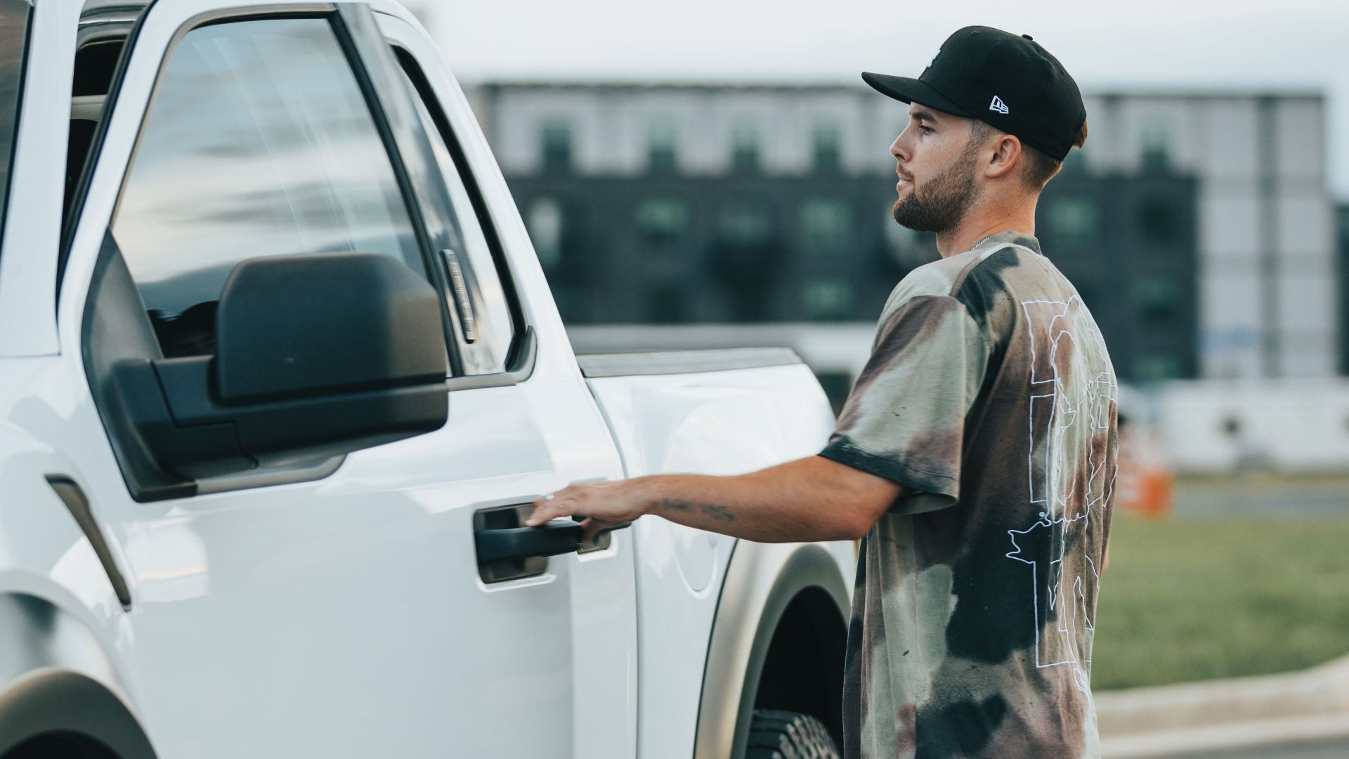 A man in casual attire opens the door of a parked white pickup truck outside.