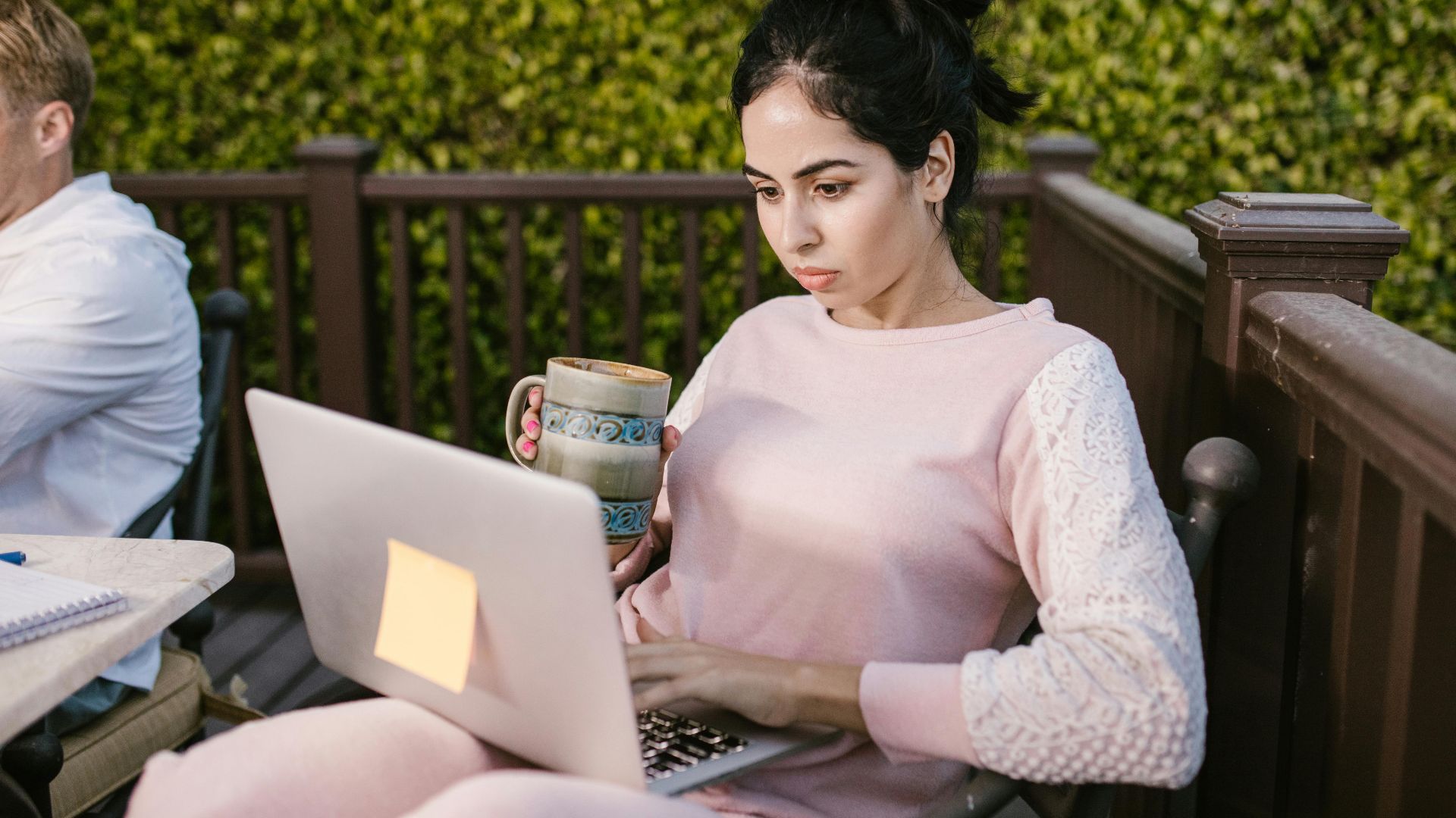 Woman focused on laptop work outdoors, holding a coffee mug. Enjoying remote work in cozy setting.