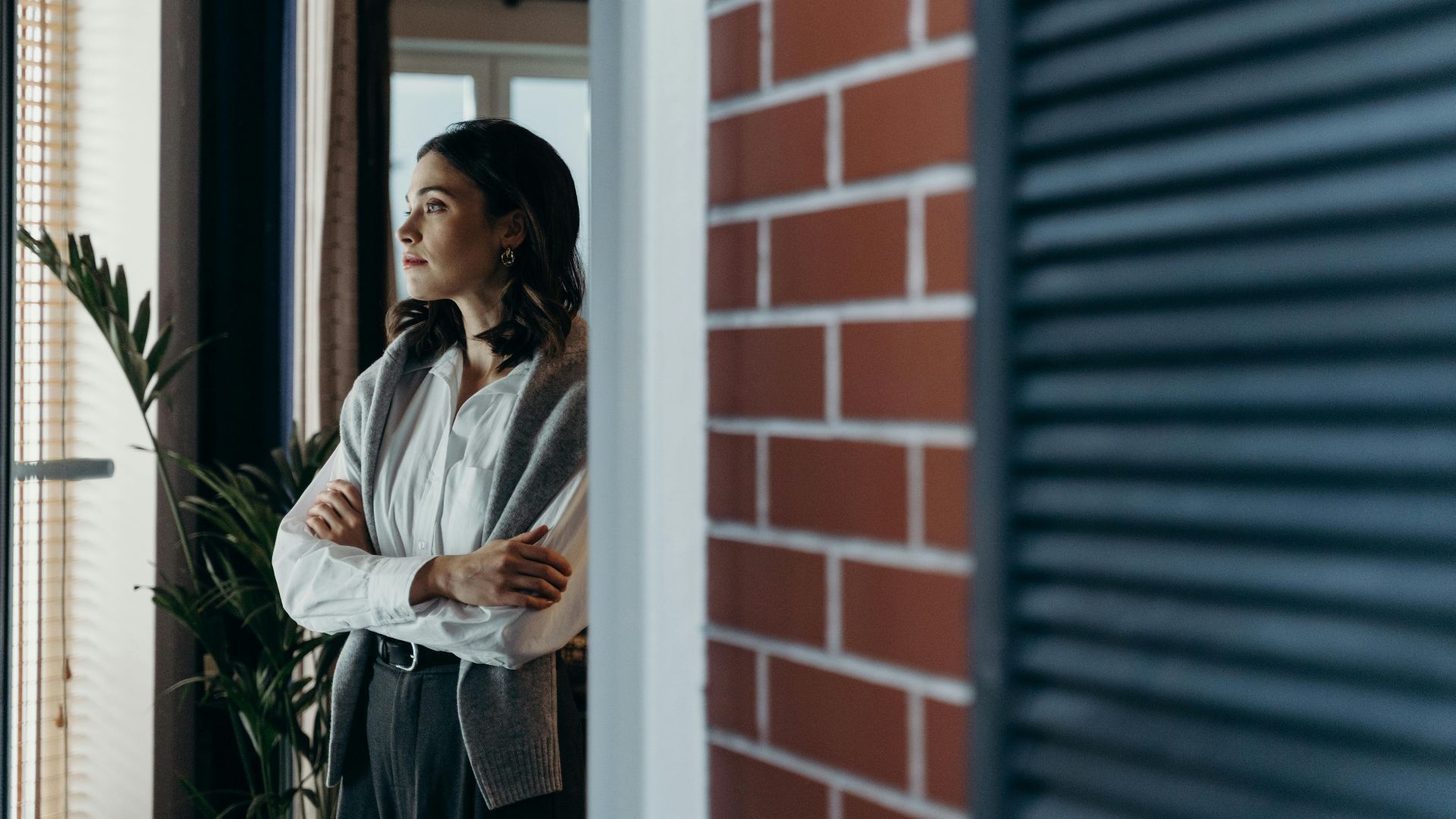 A contemplative woman stands indoors, arms crossed, gazing out the window beside a brick wall.