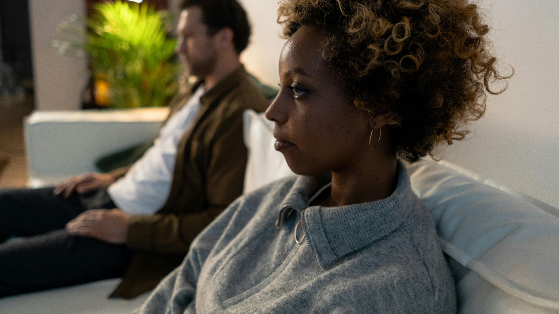 Woman with afro hair sitting pensively on a sofa indoors.