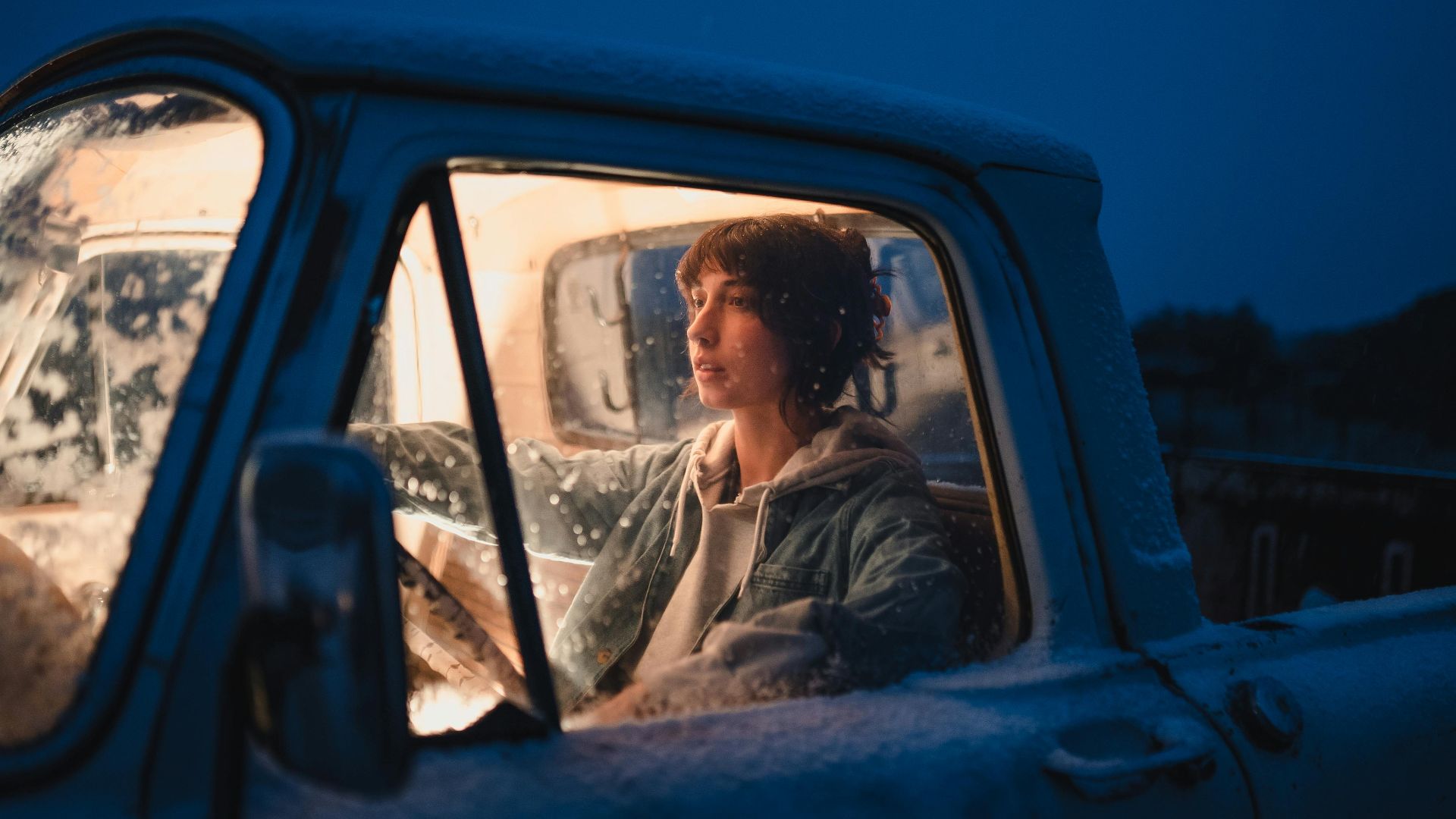 A woman sits in an illuminated, snow-covered pickup truck during a cold winter night.