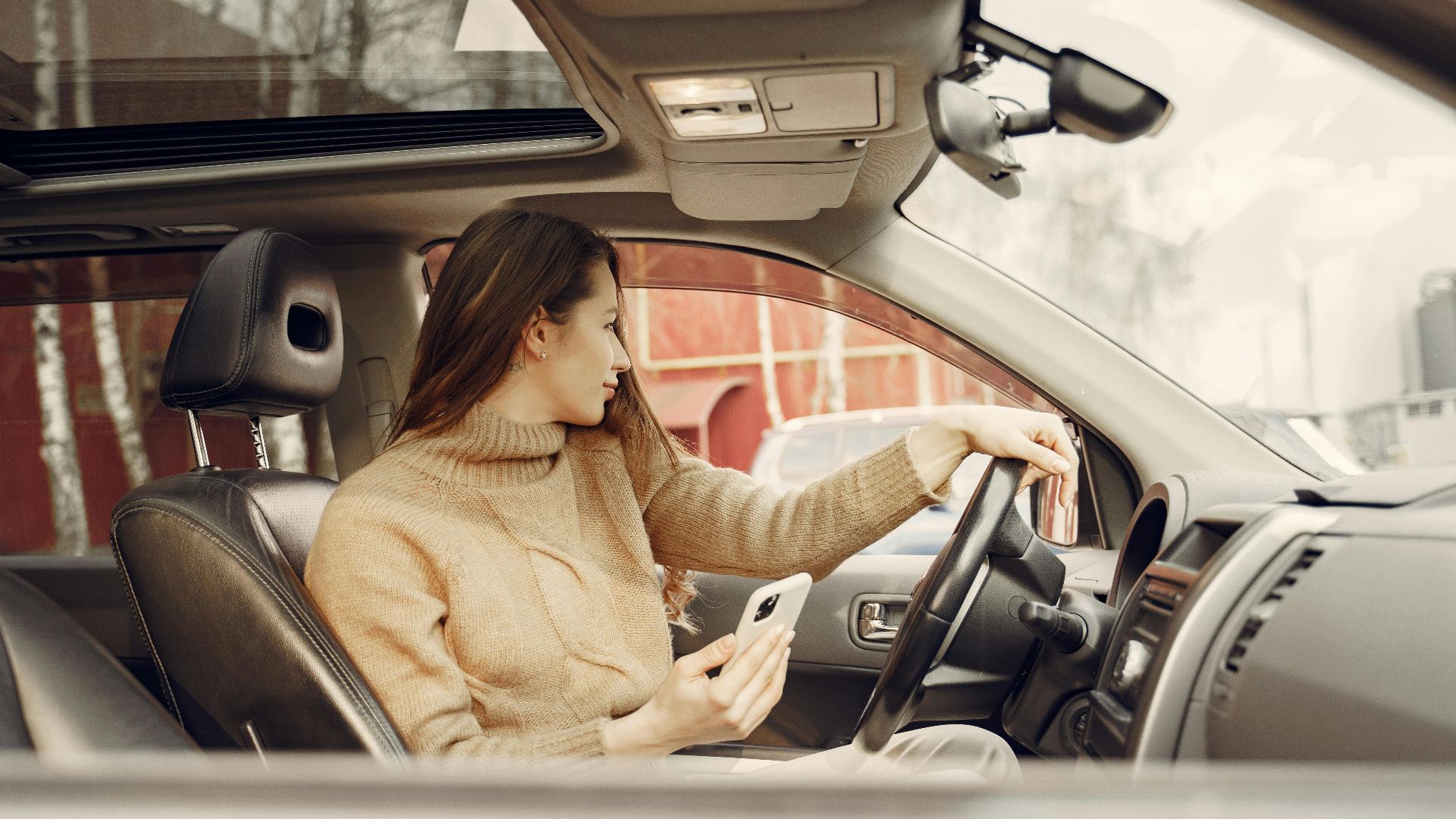 From window side view of adult woman in warm clothes sitting at wheel of modern car waiting for friend and using smartphone