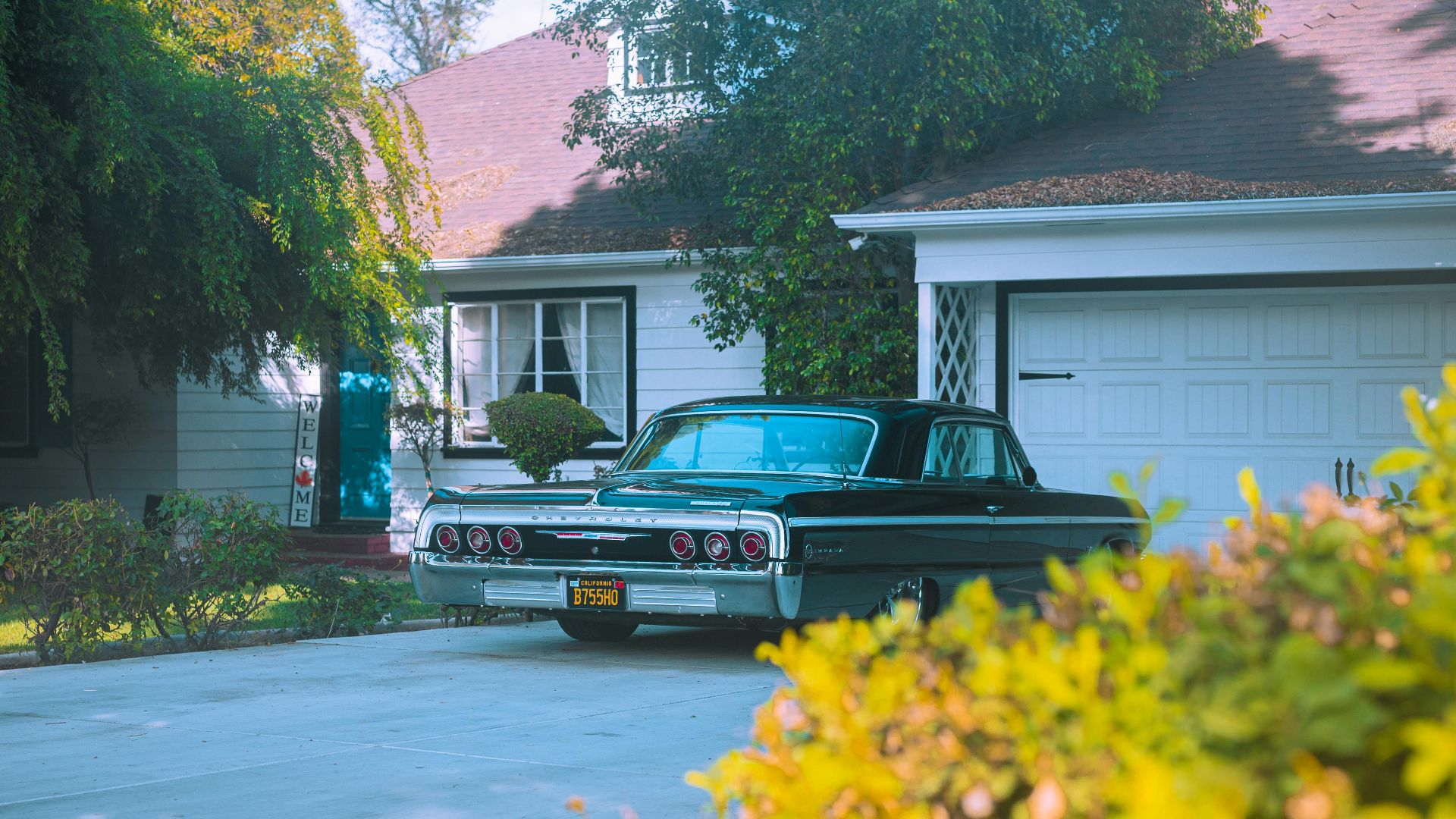 A classic black Chevrolet Impala parked in a suburban driveway surrounded by greenery.