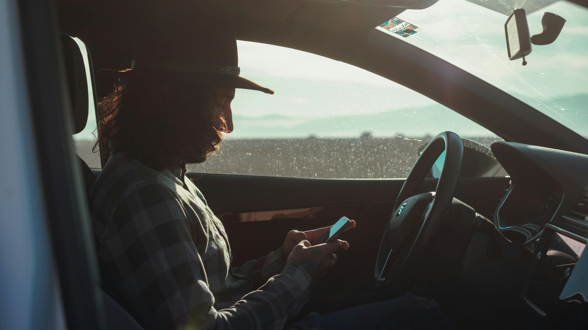 Profile of a man with a hat using a phone inside a car during the day.