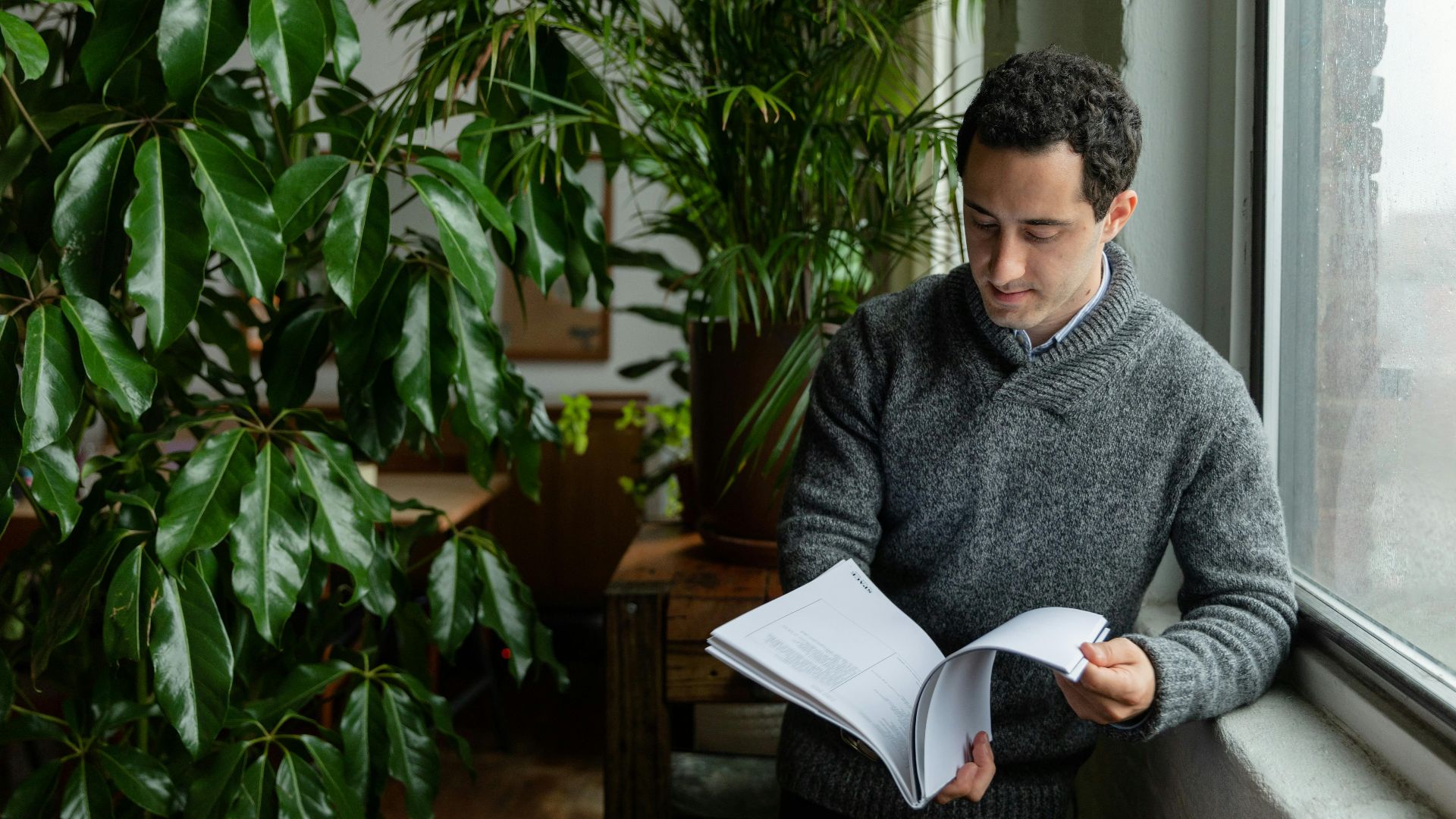 A man in a sweater reads a book next to lush indoor plants by a window.