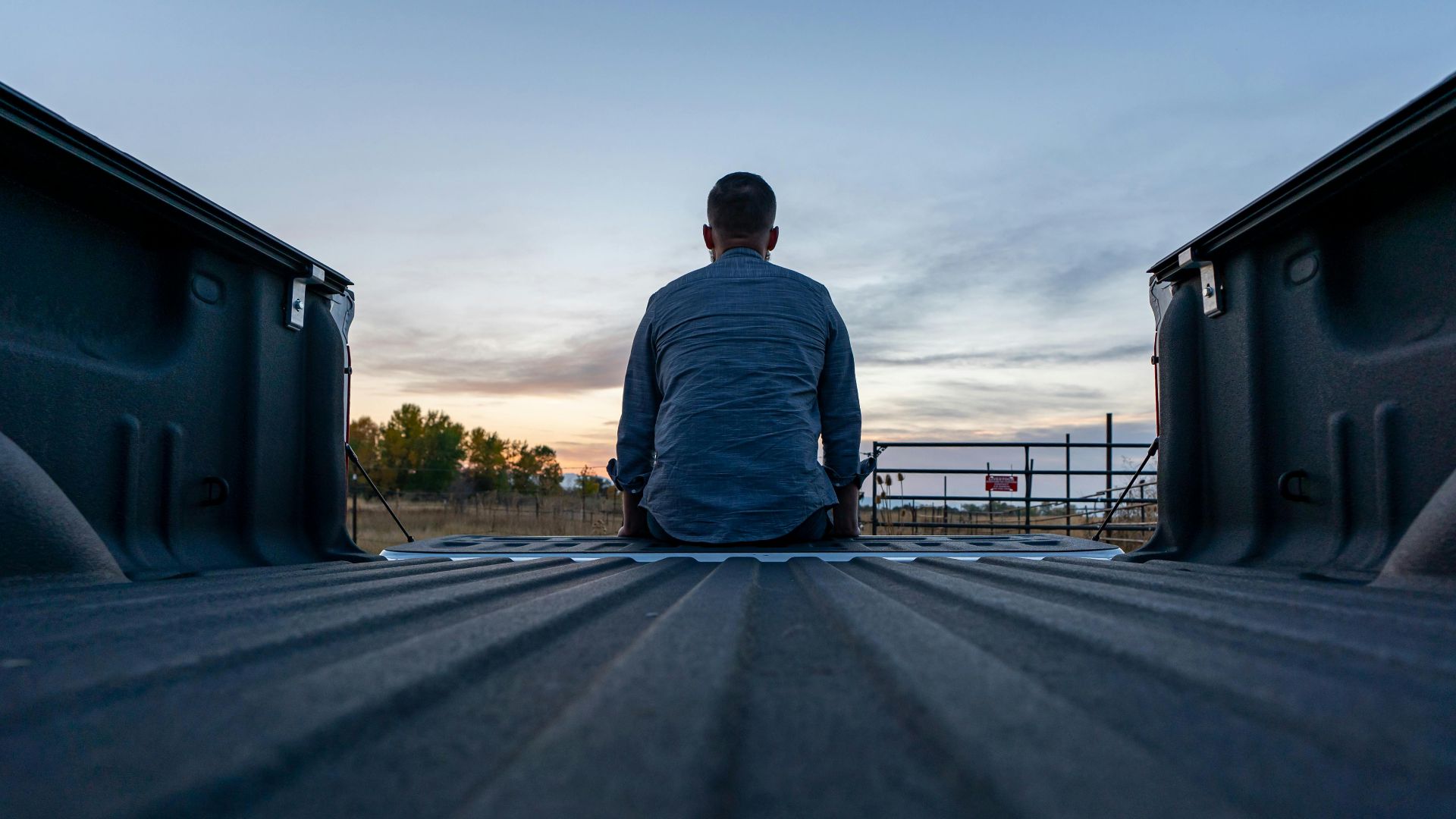 A person sits in a truck bed, facing a scenic sunset outdoors. Captured from behind.