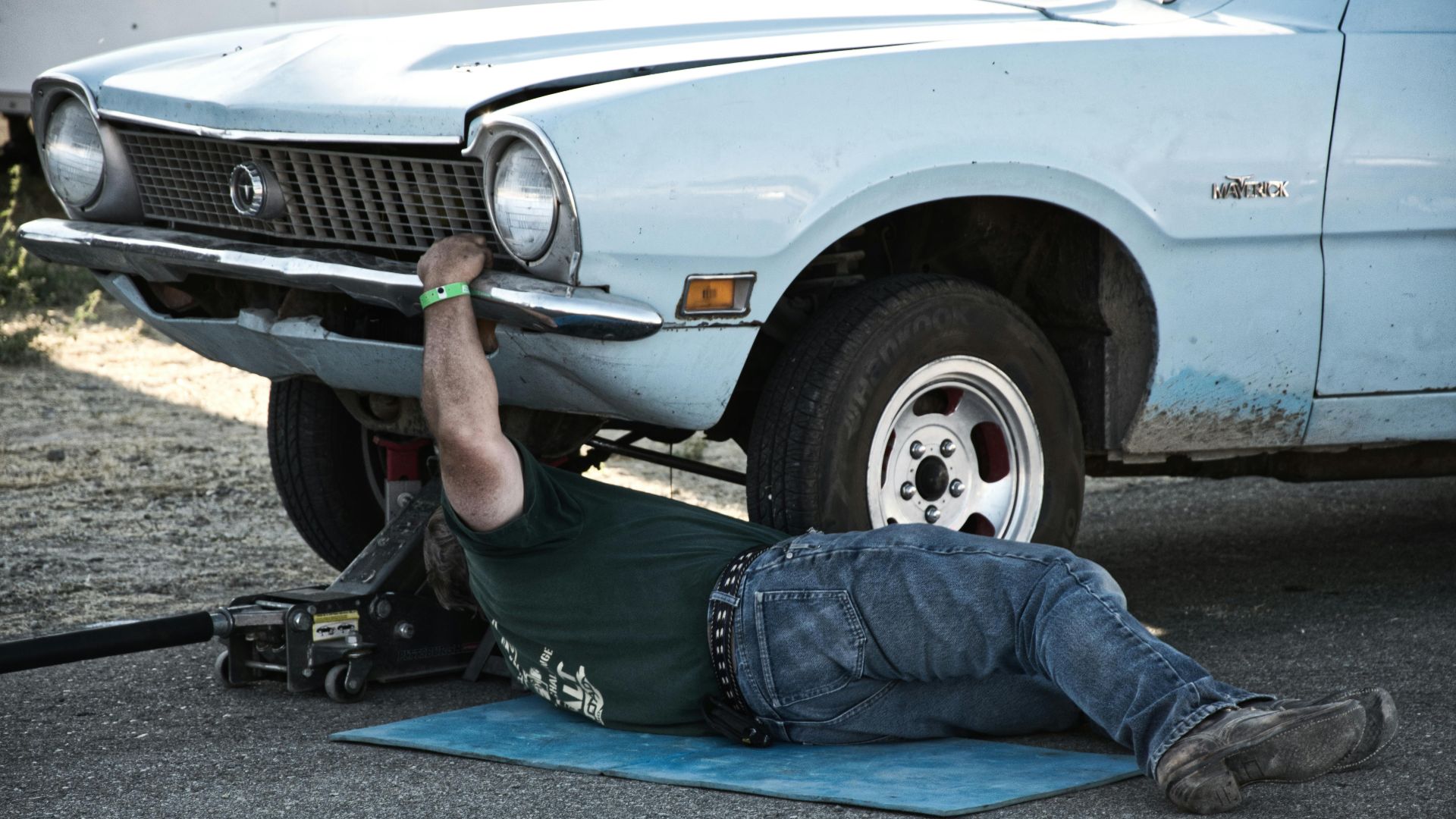 A mechanic working under a classic car on the pavement during the day.