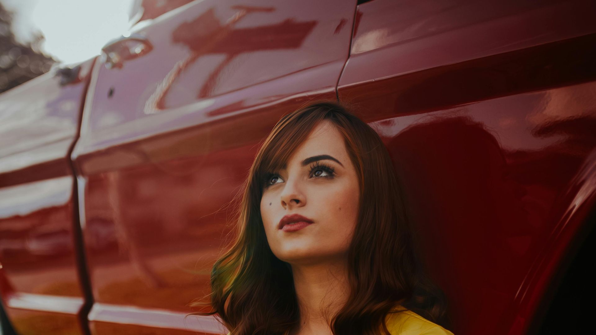 A woman with long hair sits outdoors by a vintage red pickup truck, wearing a yellow blouse.
