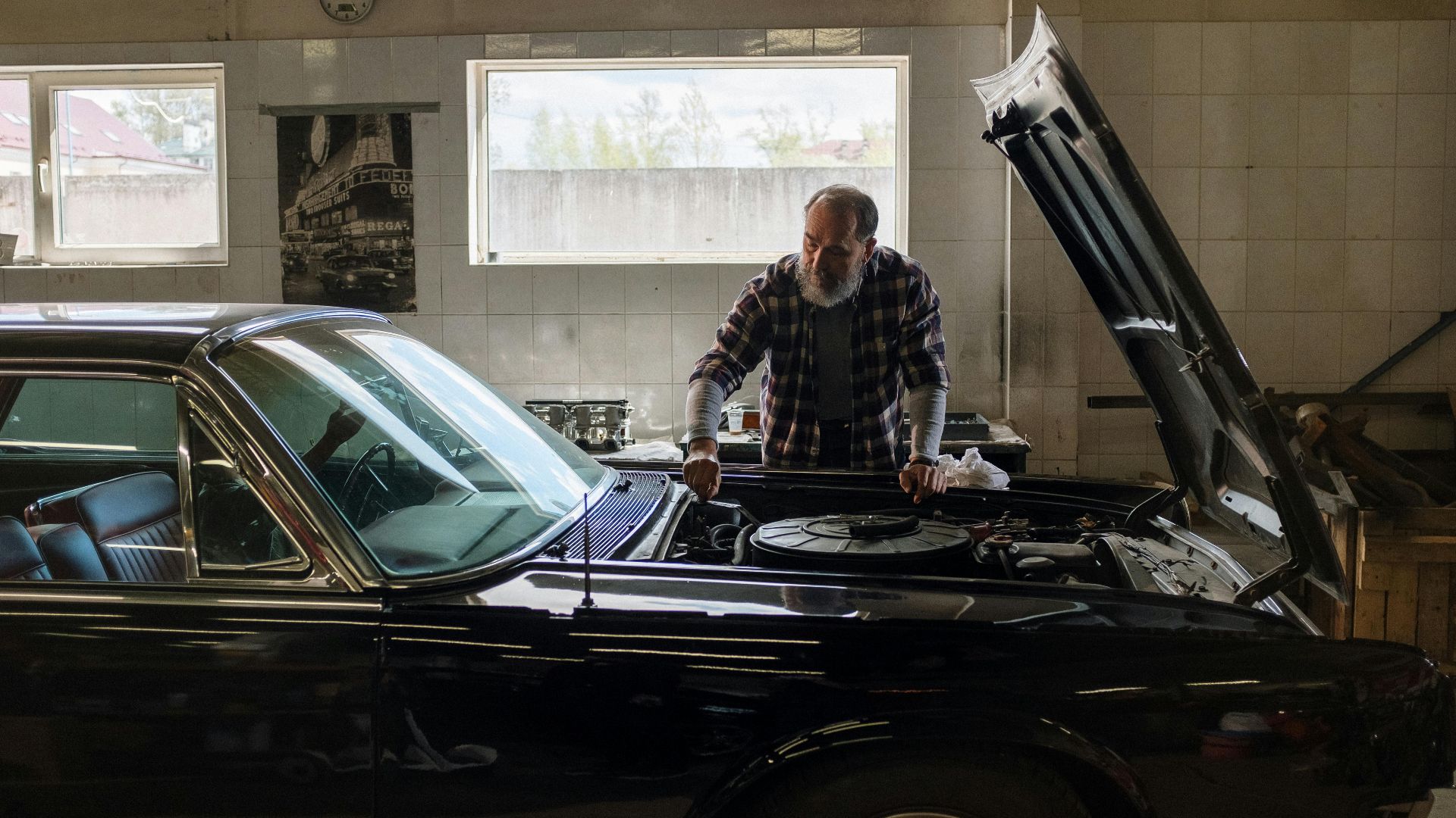 A bearded mechanic examines a classic car engine in a well-lit auto repair shop.
