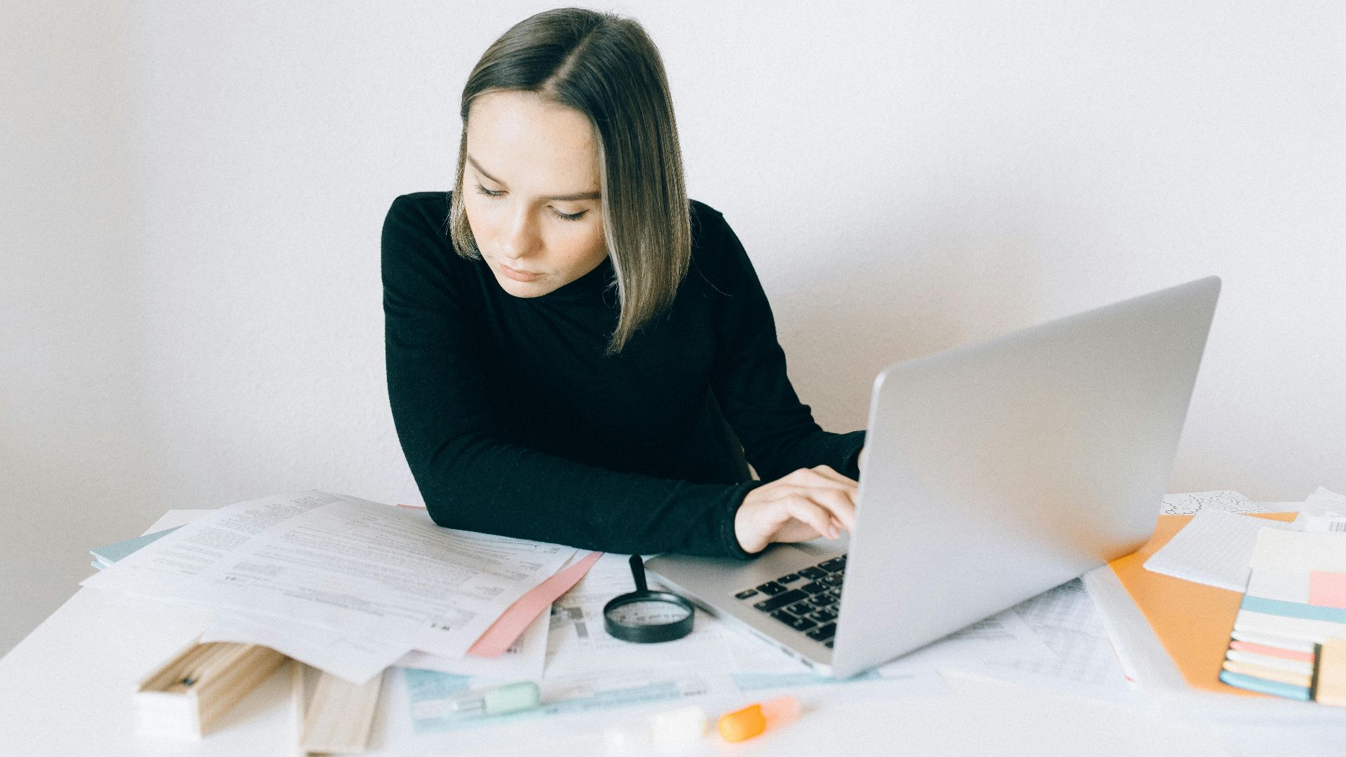 Focused woman researching on a laptop, surrounded by paperwork in a modern office.