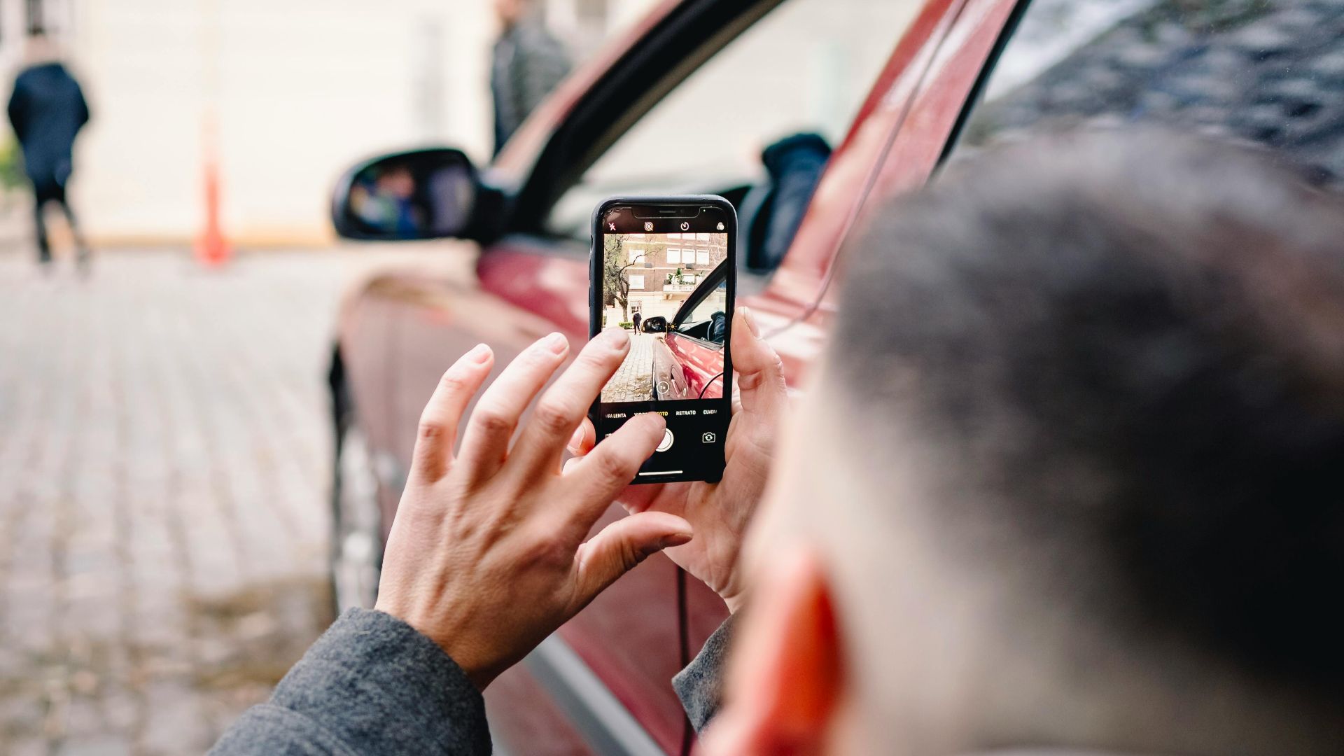 Man using smartphone to take pictures of an urban scene beside a red car.