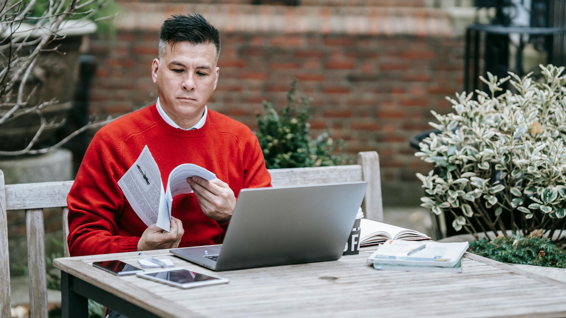 Adult male reading outside with a laptop on a wooden table, embracing remote work lifestyle.