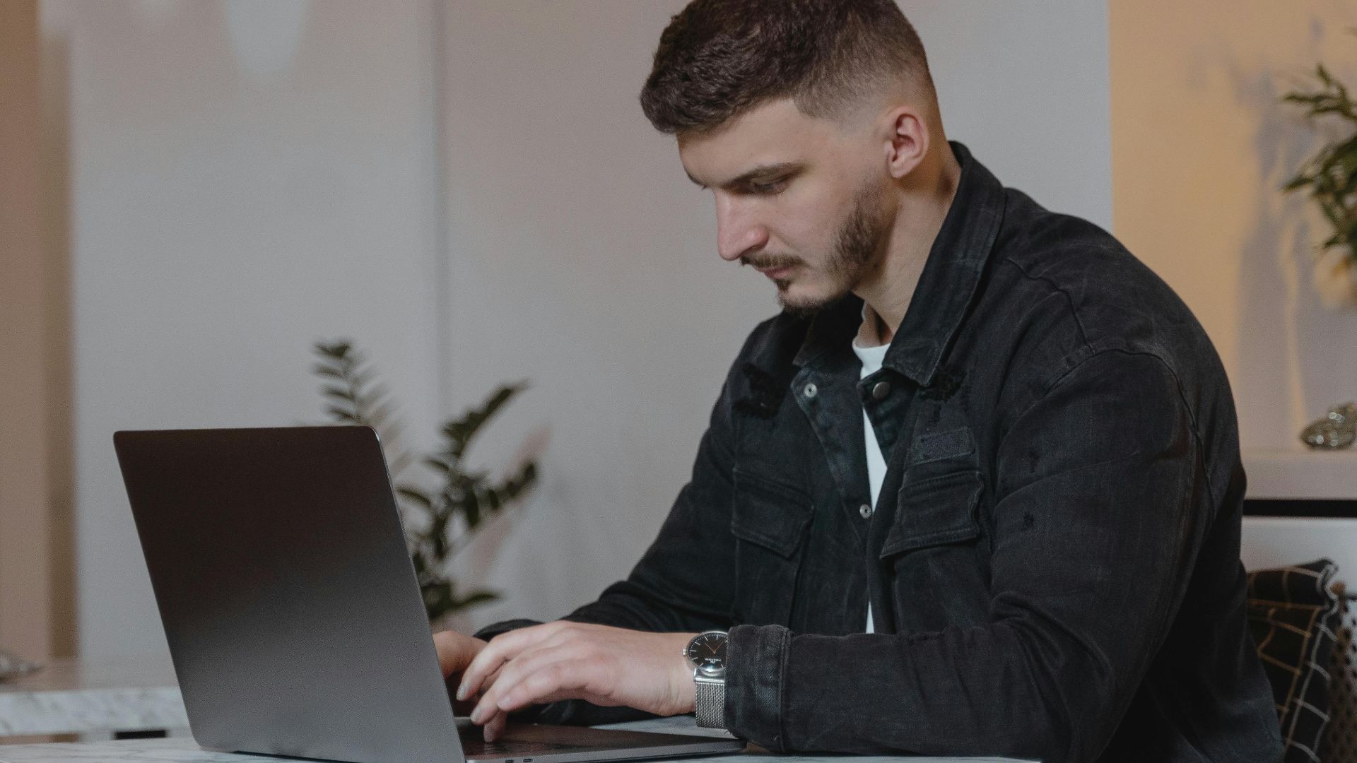 Young man in a denim jacket working on a laptop indoors, showcasing modern remote work lifestyle.
