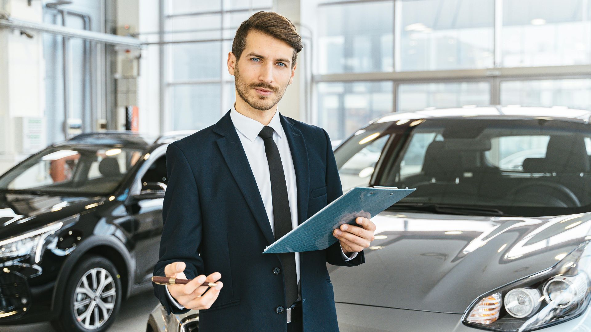 Professional car dealer in business suit holding clipboard in a bright car showroom.