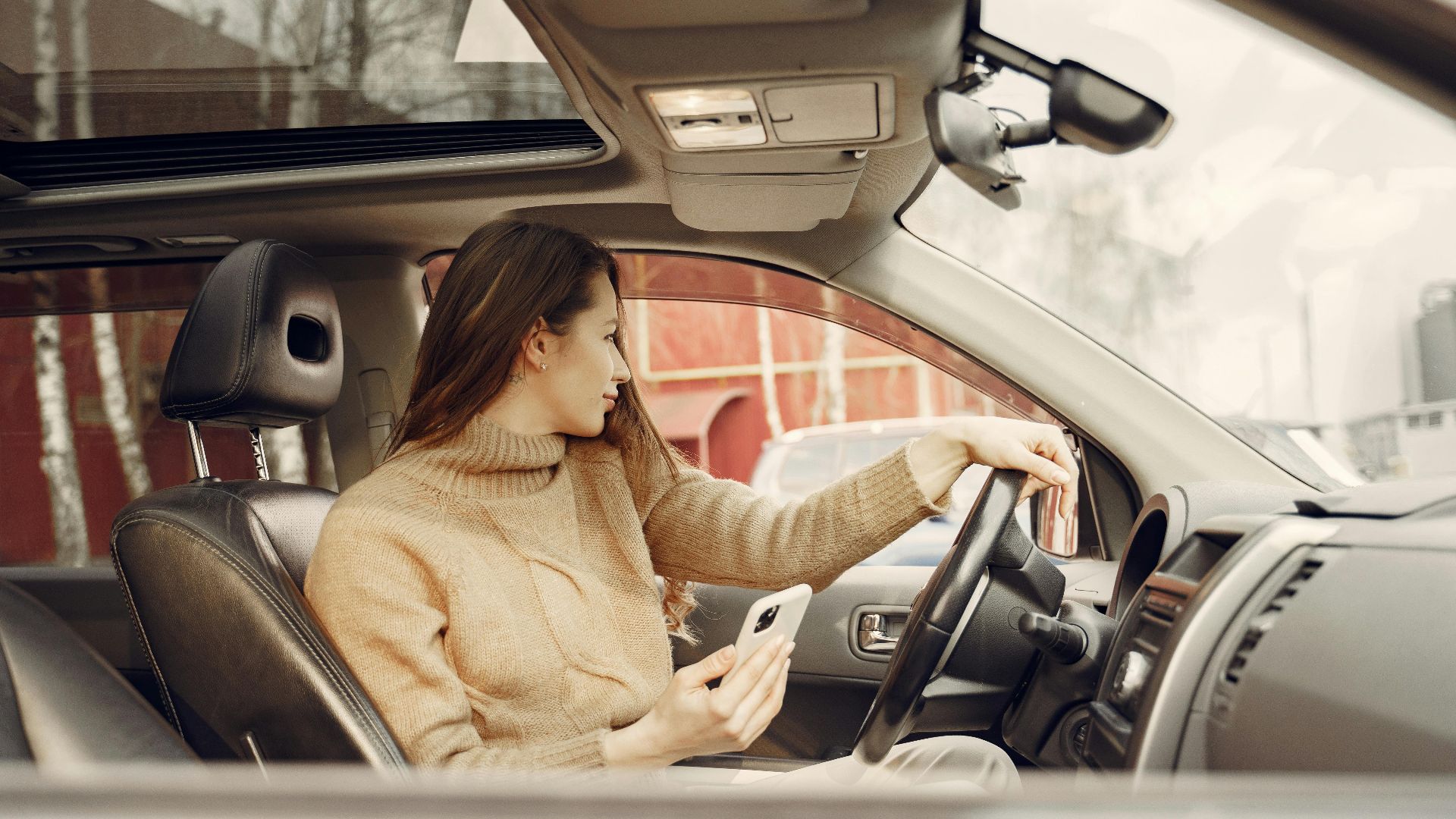 From window side view of adult woman in warm clothes sitting at wheel of modern car waiting for friend and using smartphone