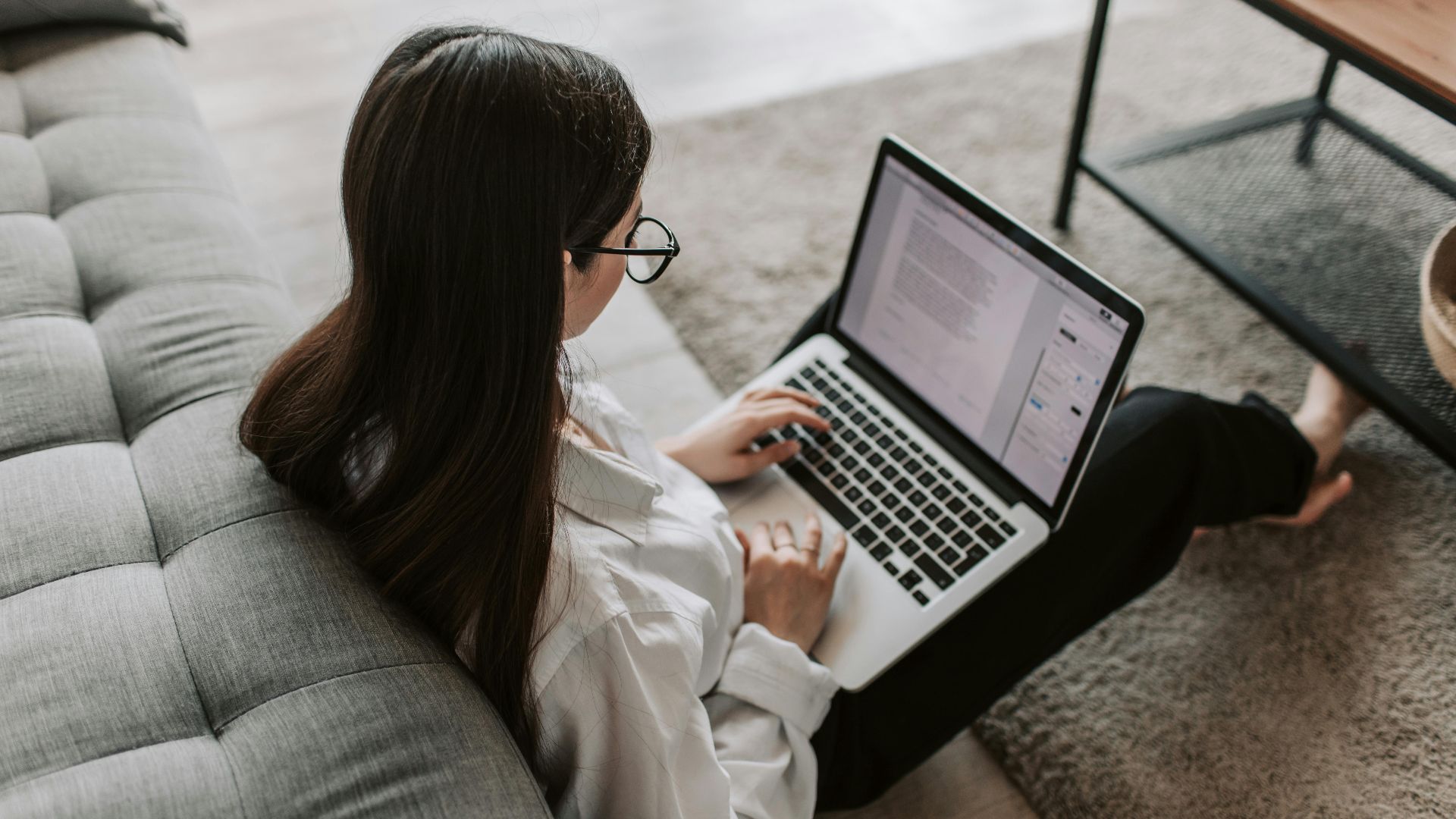 Woman working remotely with a laptop on the floor next to a sofa, enjoying comfortable home office setup.