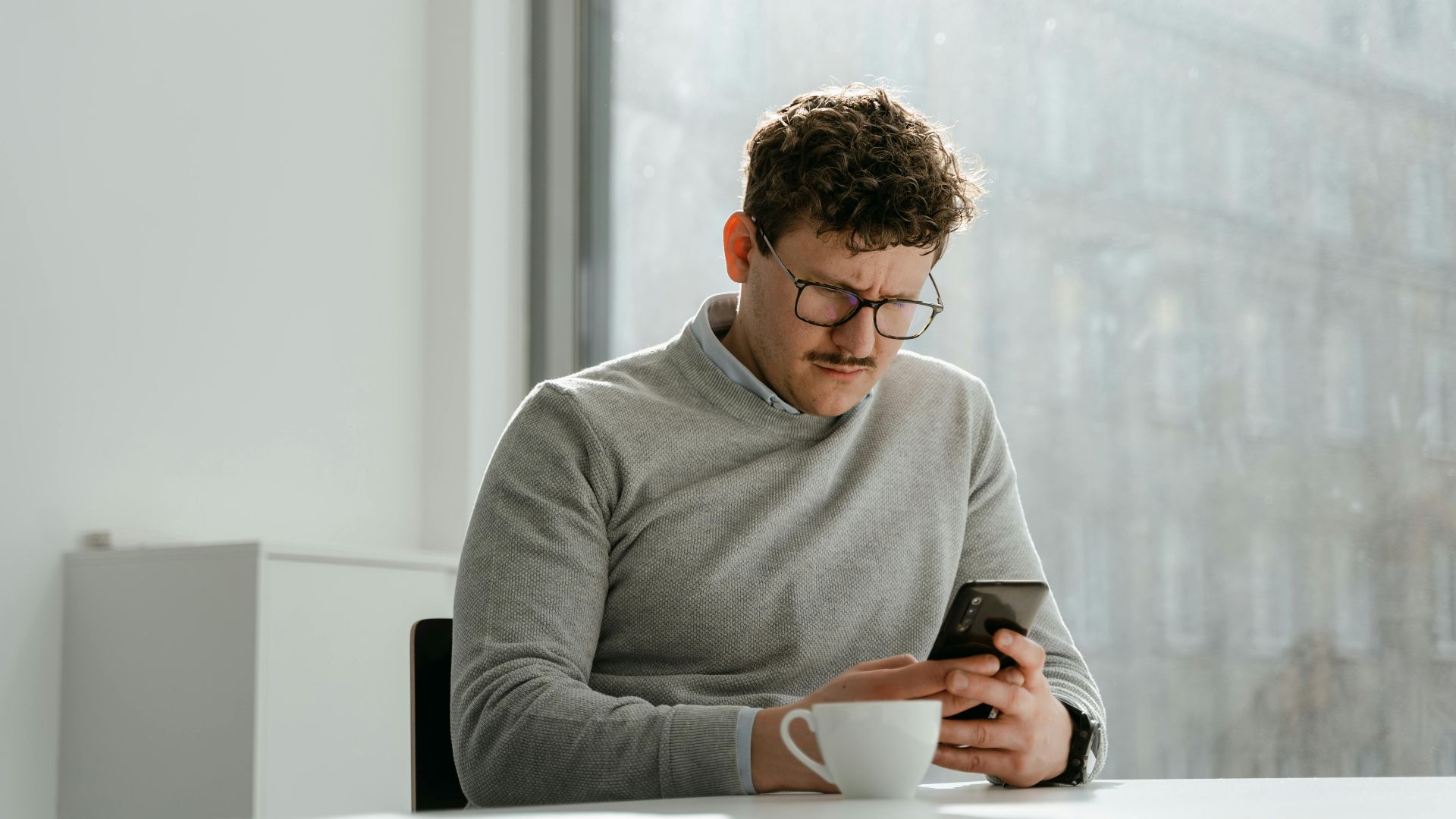 Adult man wearing glasses using smartphone in sunny office space, holding a mug.