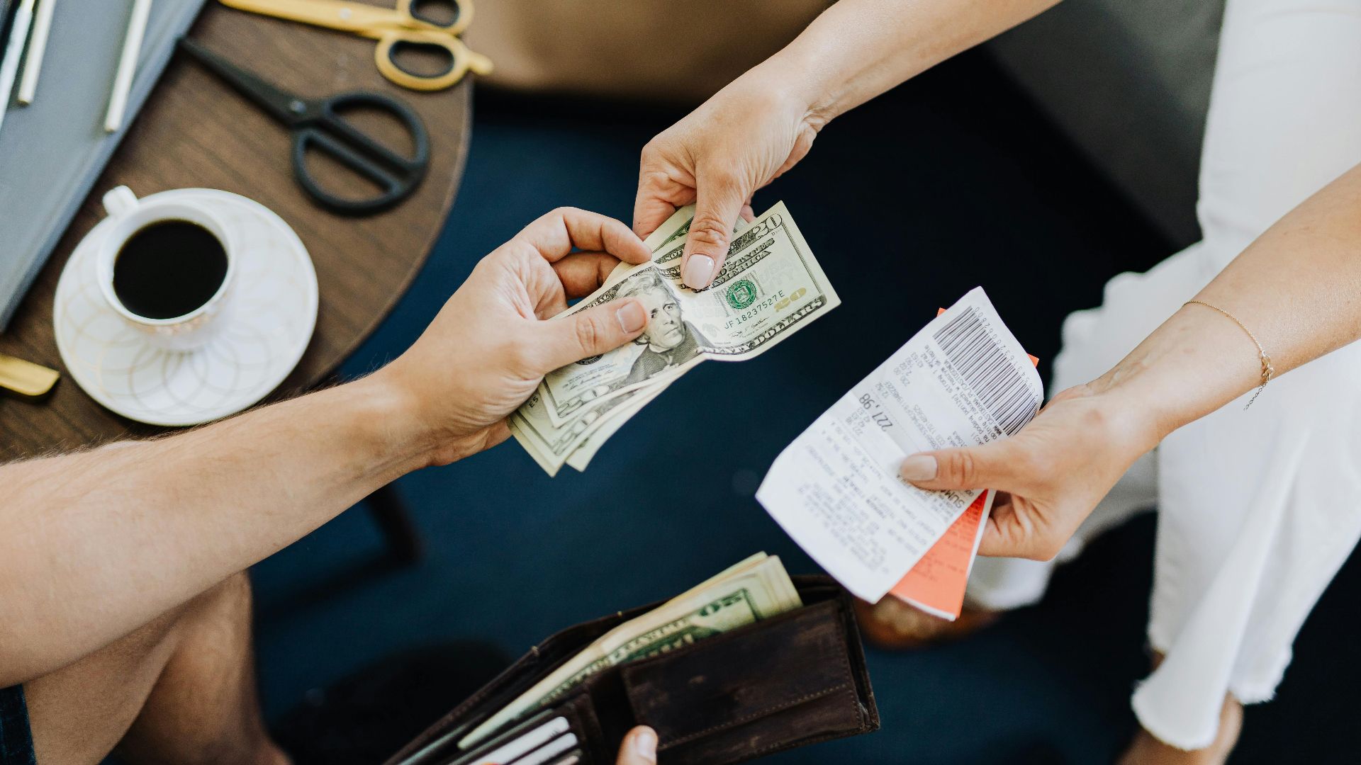 Close-up of a transaction with US dollar bills and receipts being exchanged over a table with coffee and scissors.