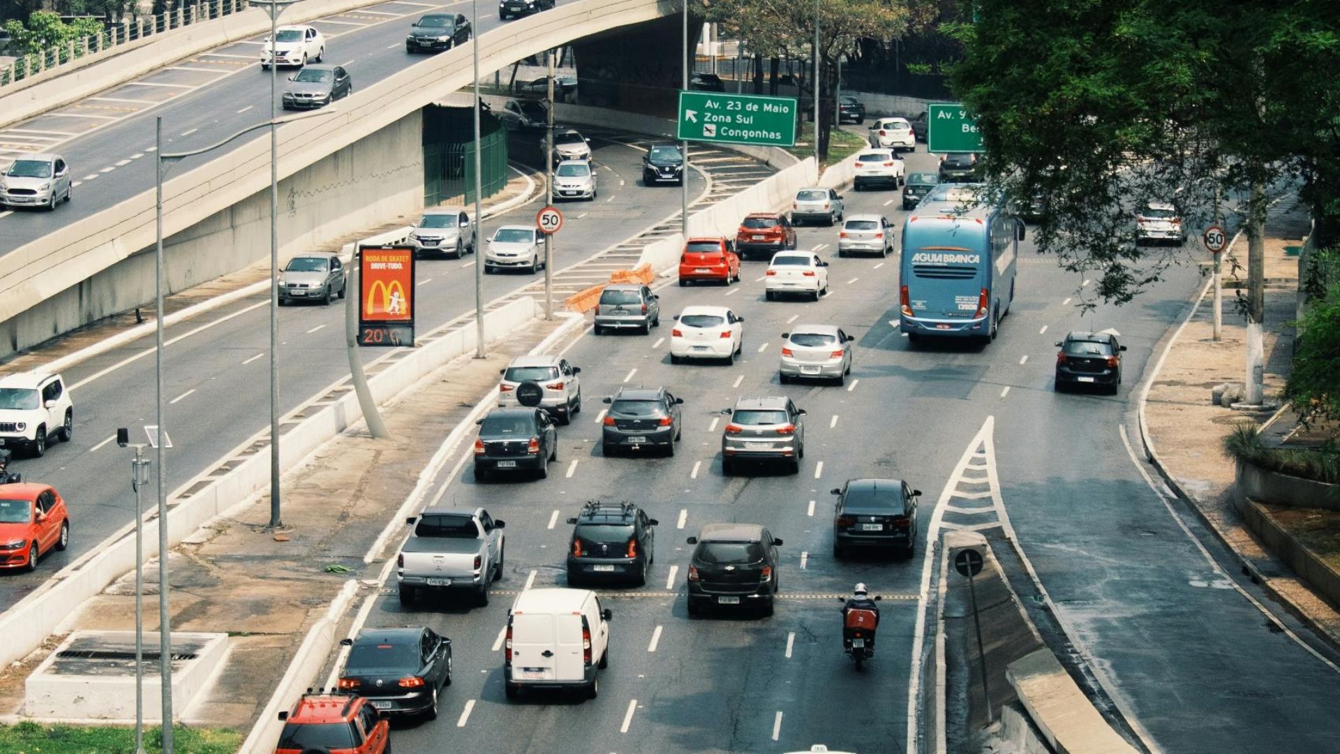 Cars and buses navigate a busy city road surrounded by tall buildings and lush greenery.