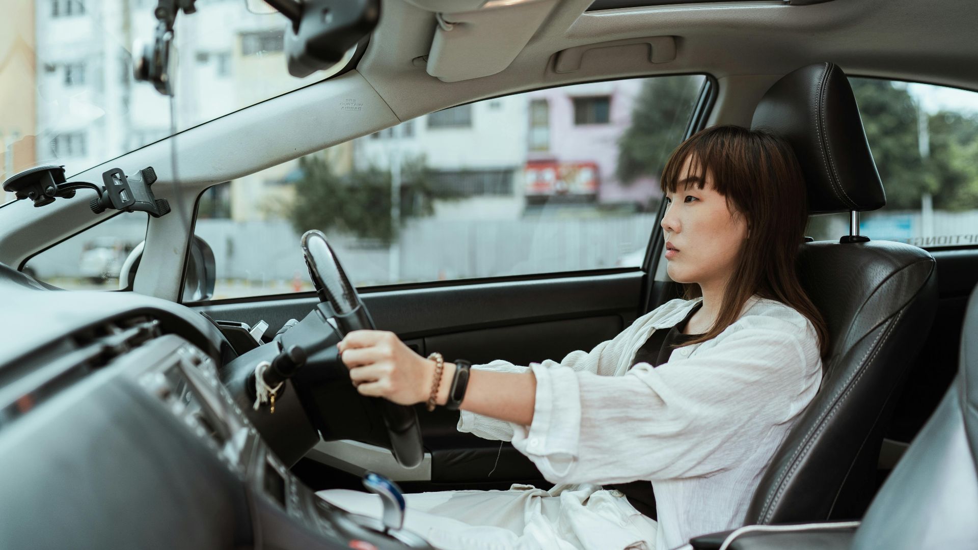 Side view unemotional Asian female in casual white clothes steering right hand drive car along urban streets in daytime