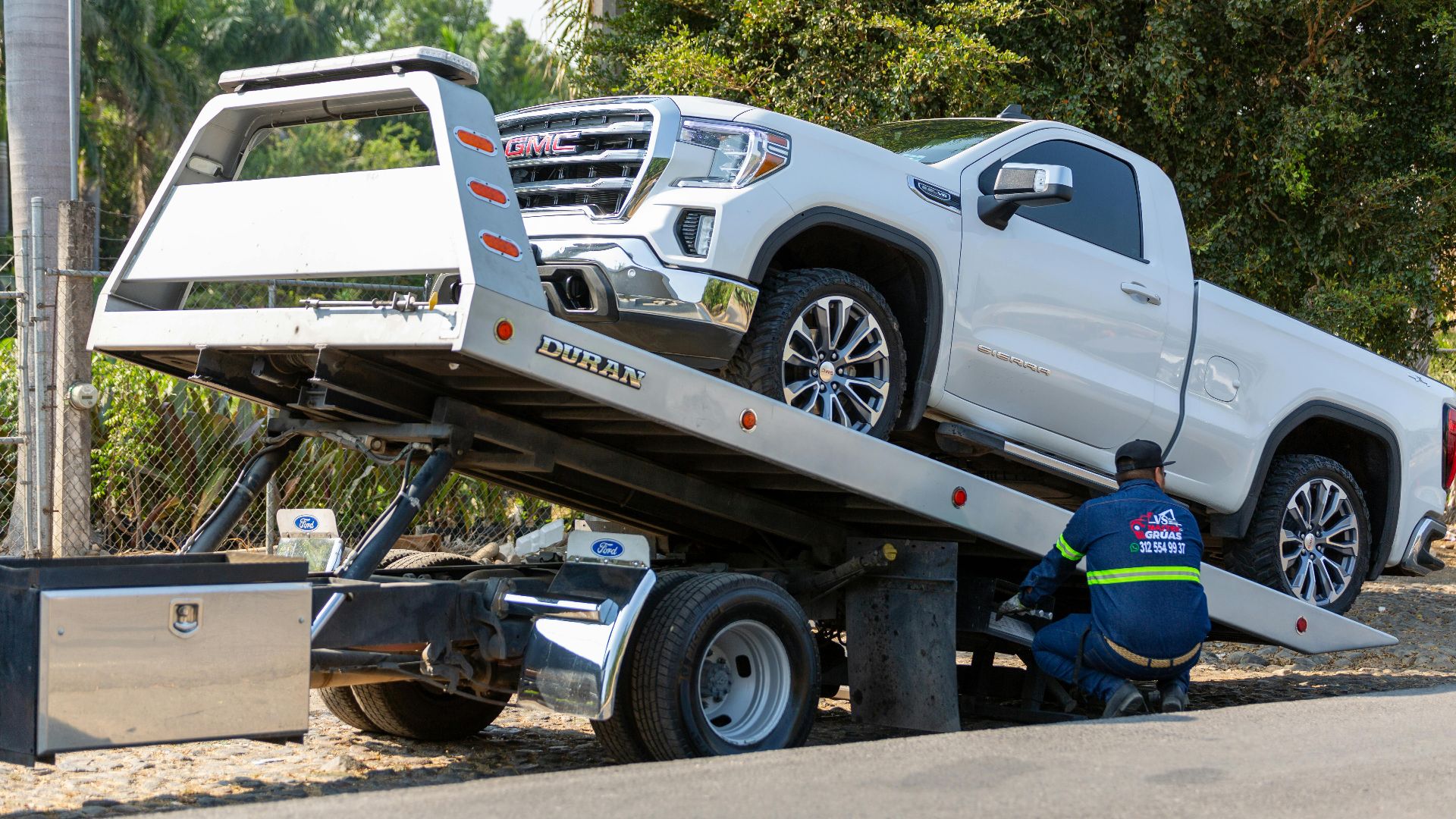 Tow truck operator loading white GMC pickup truck on street in daytime.