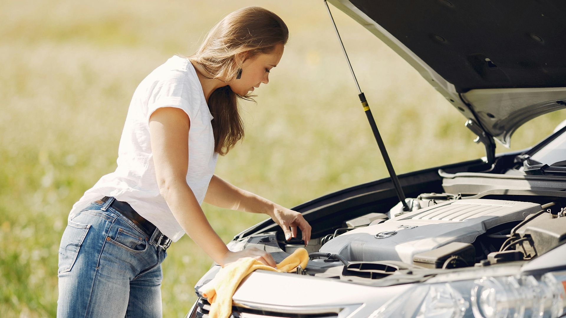 Side view of female driver in white t shirt and jeans checking damage of car engine and wiping details under open hood while standing against green field in summer day