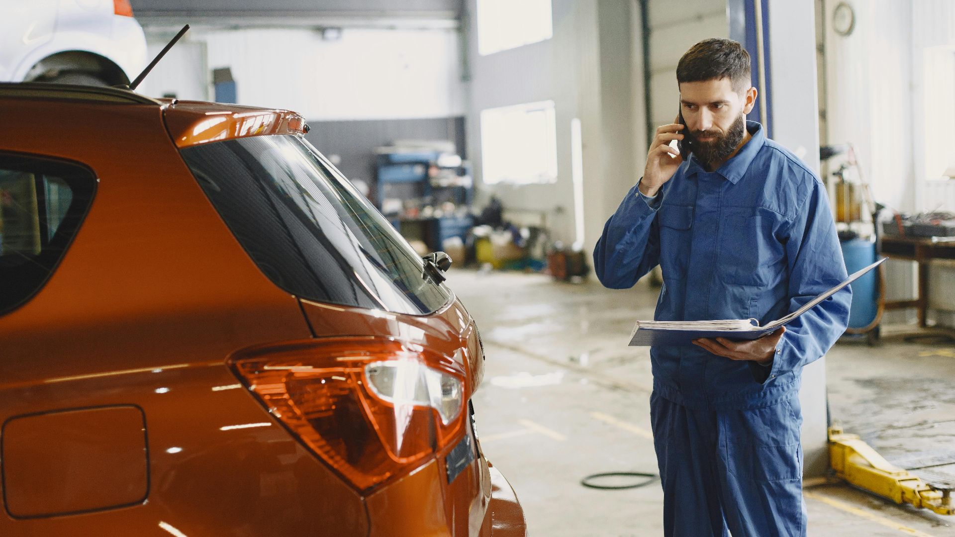 Mechanic in a blue uniform talks on phone beside a red car in an auto repair shop.