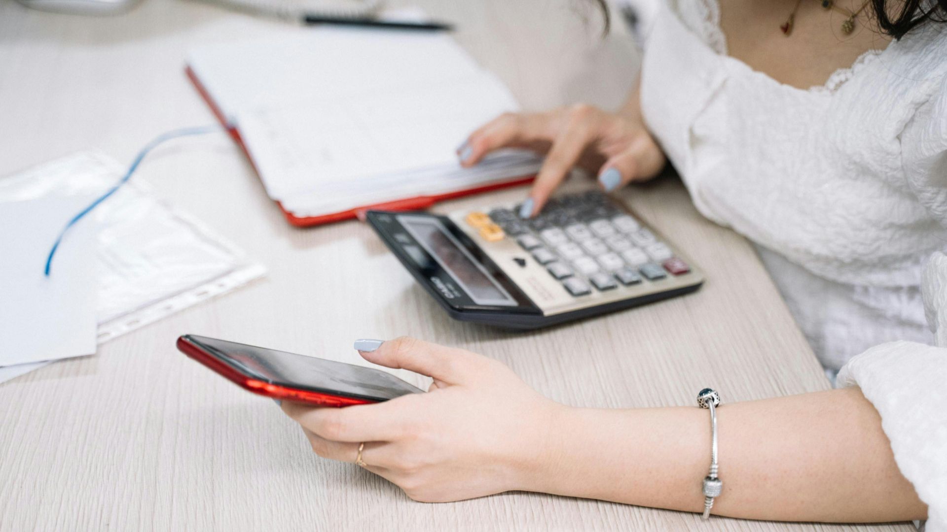 A woman managing finances using a calculator and smartphone at her desk, showcasing multitasking in an office setting.
